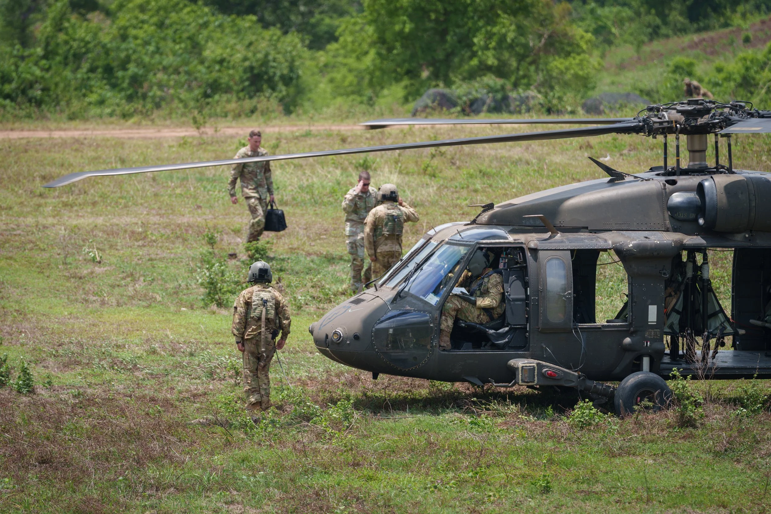 Military personnel preparing a black military helicopter on a grassy field, with trees and rocks in the background.