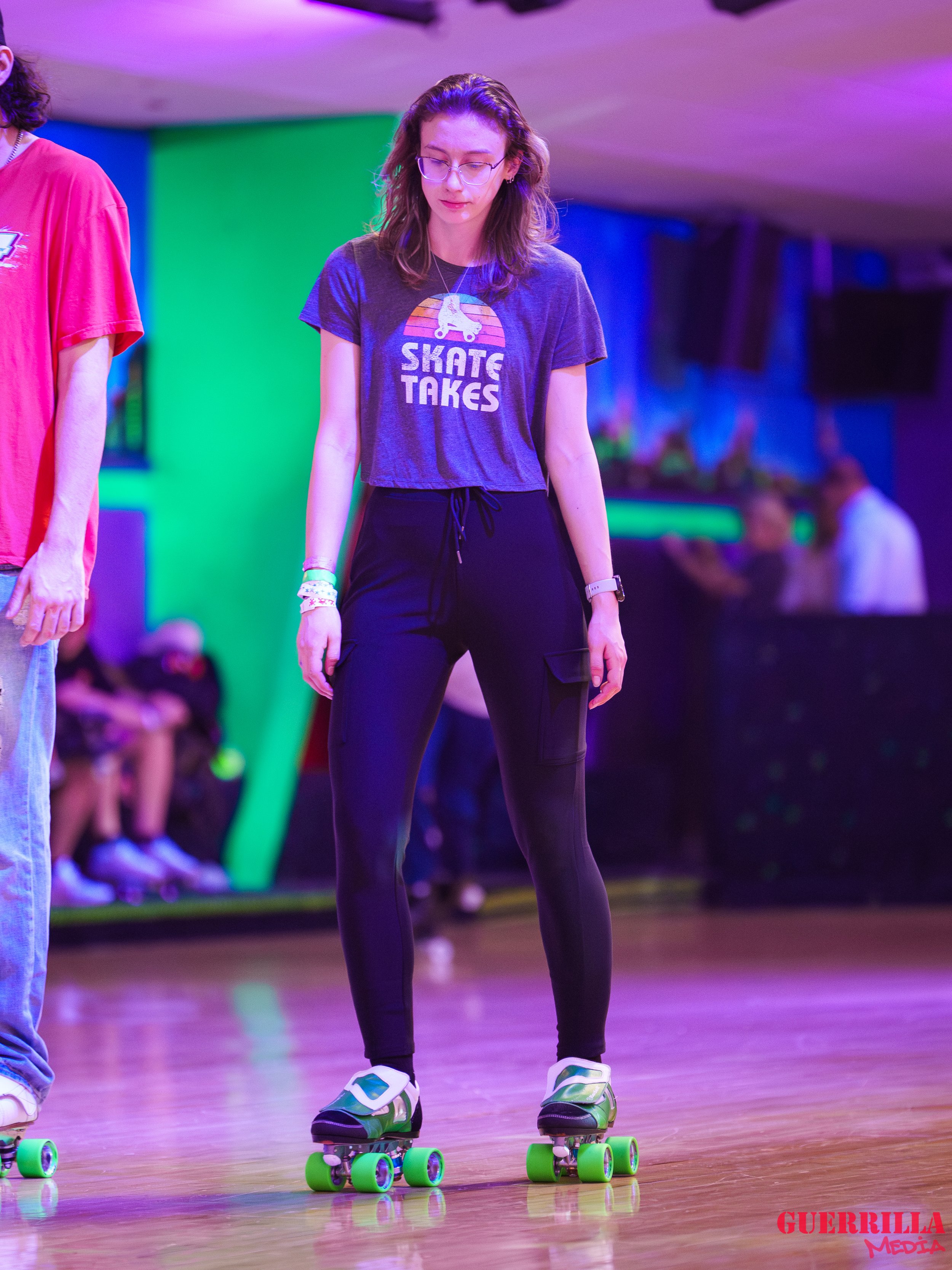 Young woman wearing glasses and a t-shirt that says 'Skate Takes,' standing on a green roller skate, on a wooden floor with colorful lighting, in an indoor skating rink or entertainment venue.