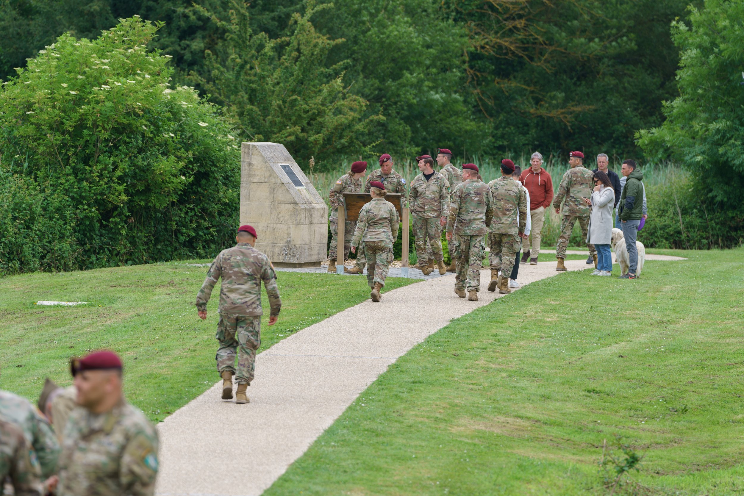 Military personnel walking along a park pathway, some in camouflage uniforms and maroon berets, others in civilian clothing, with a woman and dog on the right.