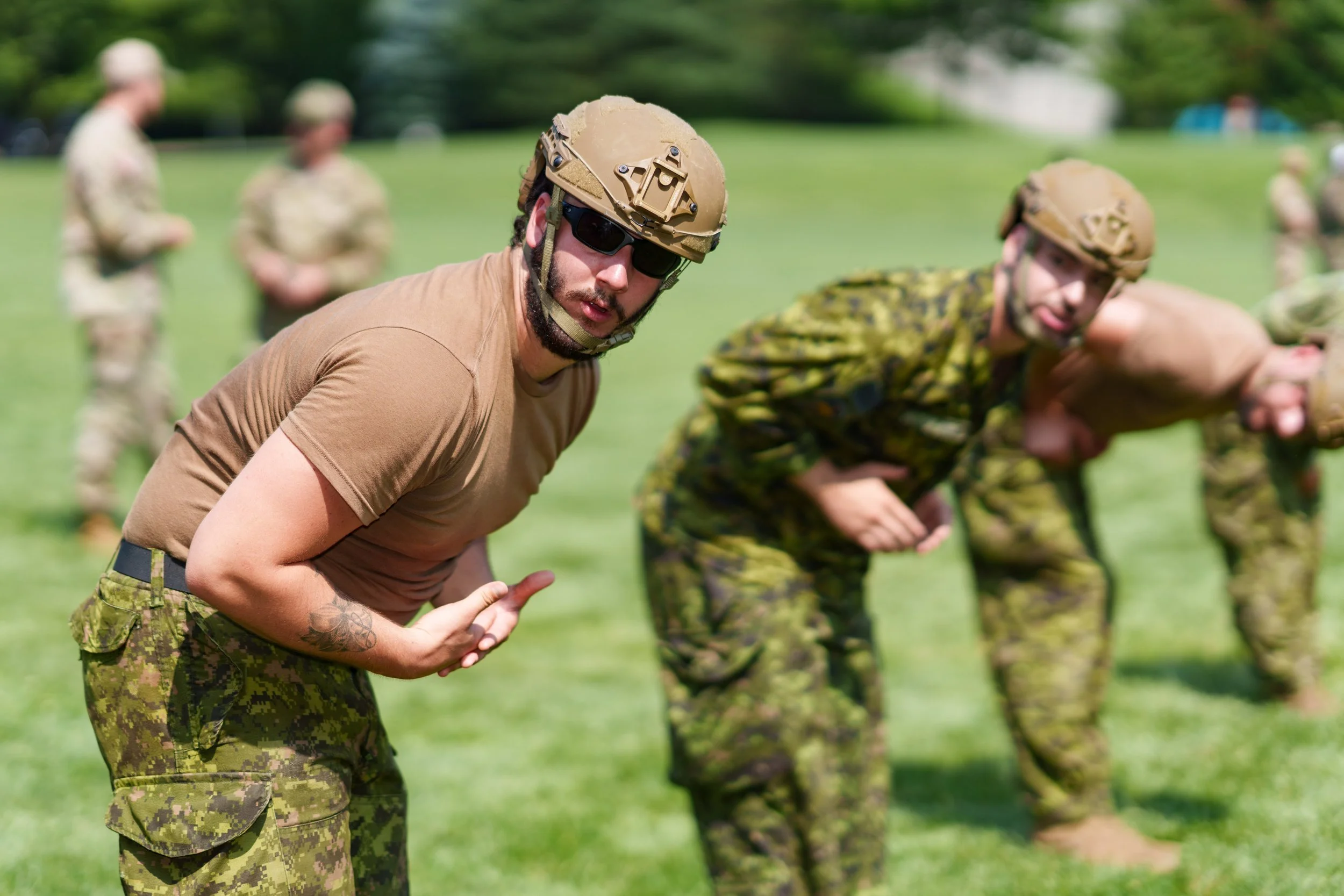 Military personnel in camouflage and tan uniforms participating in training exercises on a grassy field, with some wearing helmets and sunglasses, in an outdoor setting.