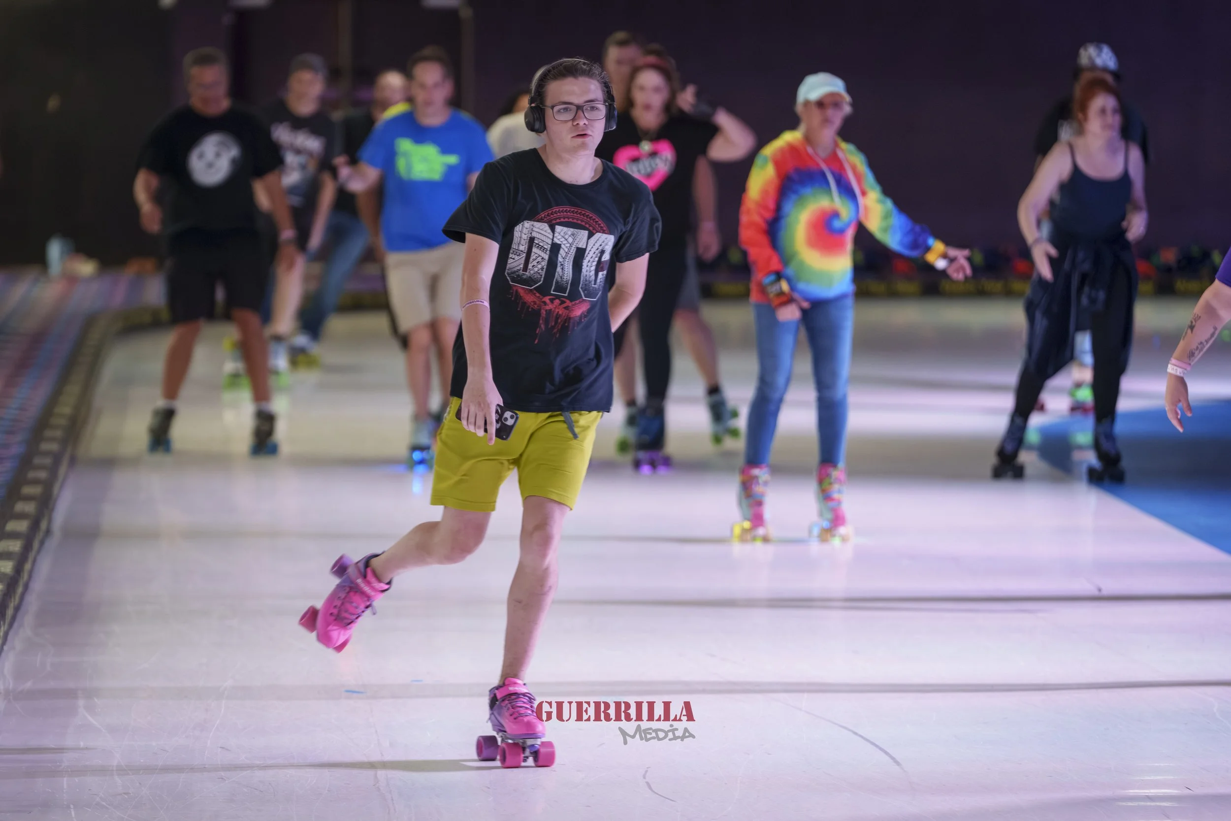 People roller skating indoors, with a woman in front wearing yellow shorts, a black T-shirt, pink roller skates, and headphones, holding a phone, and other skaters in colorful outfits in the background.