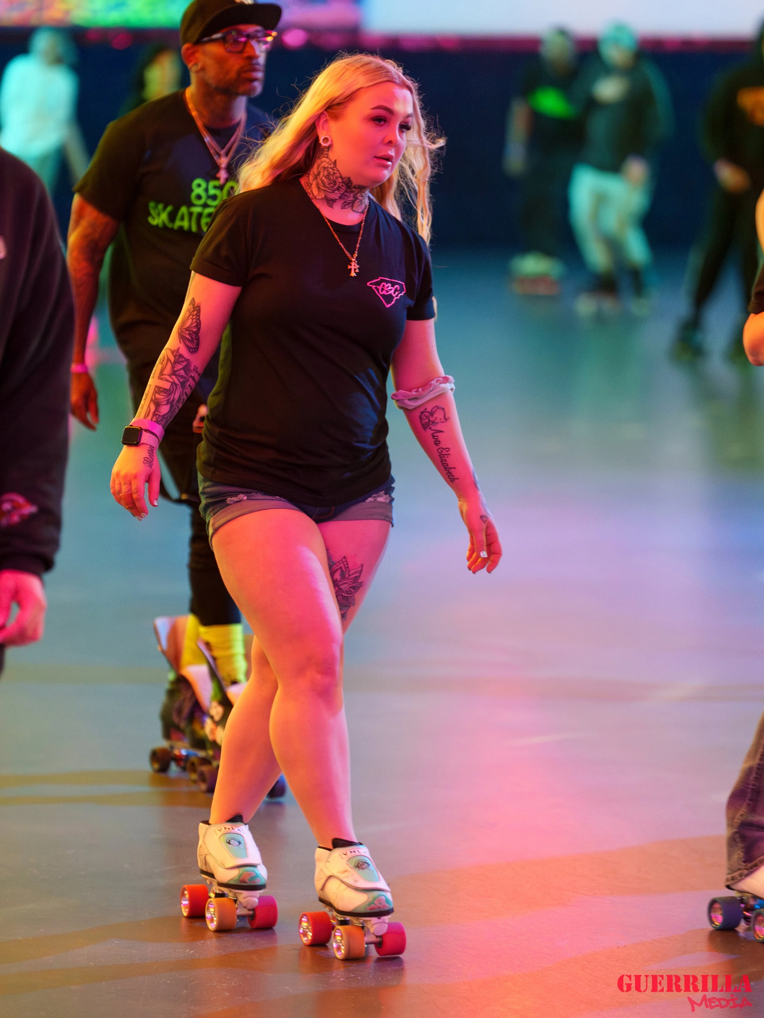A woman roller skating indoors with other skaters in the background, wearing a black T-shirt, shorts, and tattoos on her arms and legs.