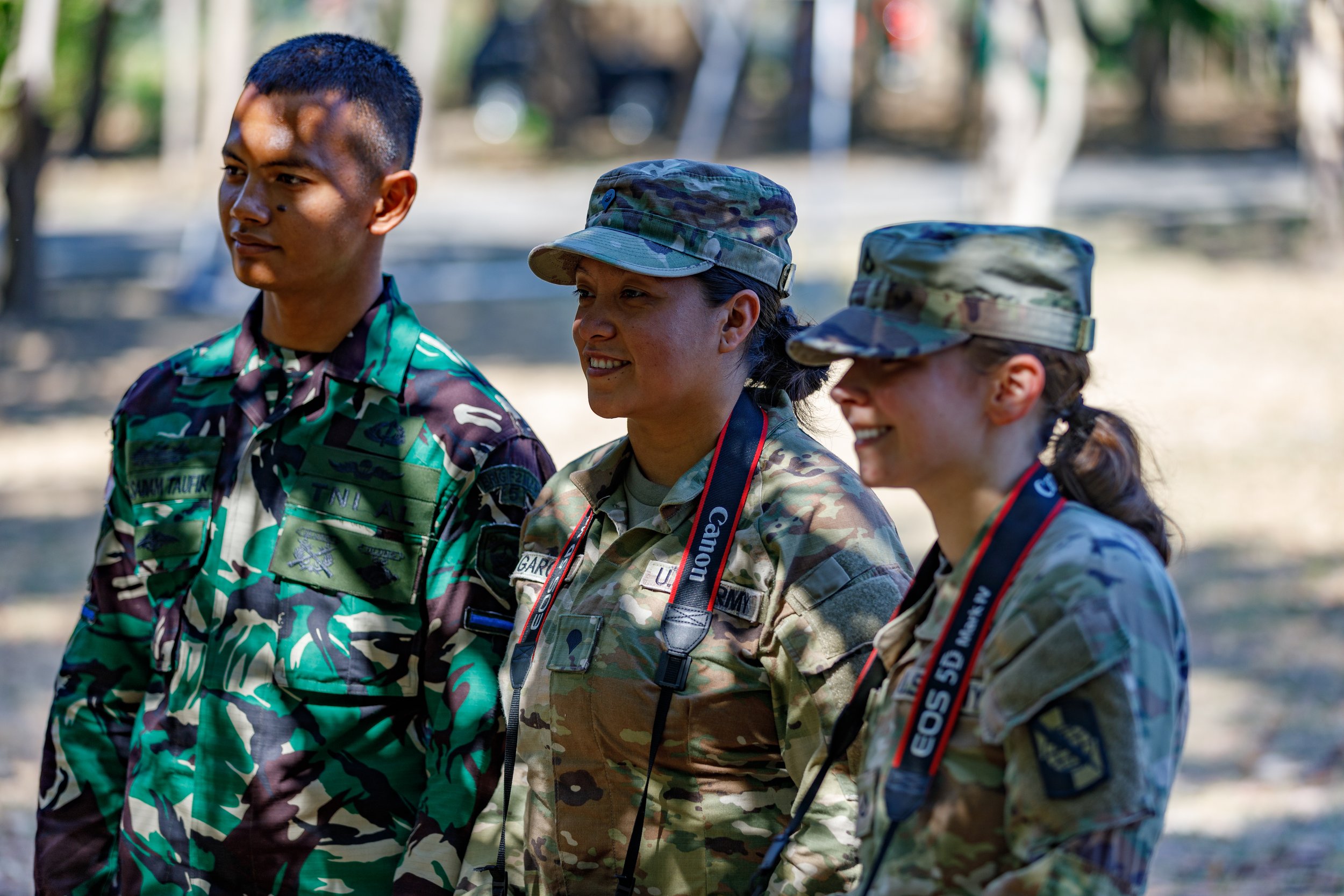Three women in military camouflage uniforms standing outdoors during daytime, smiling, with two carrying Canon cameras around their necks.