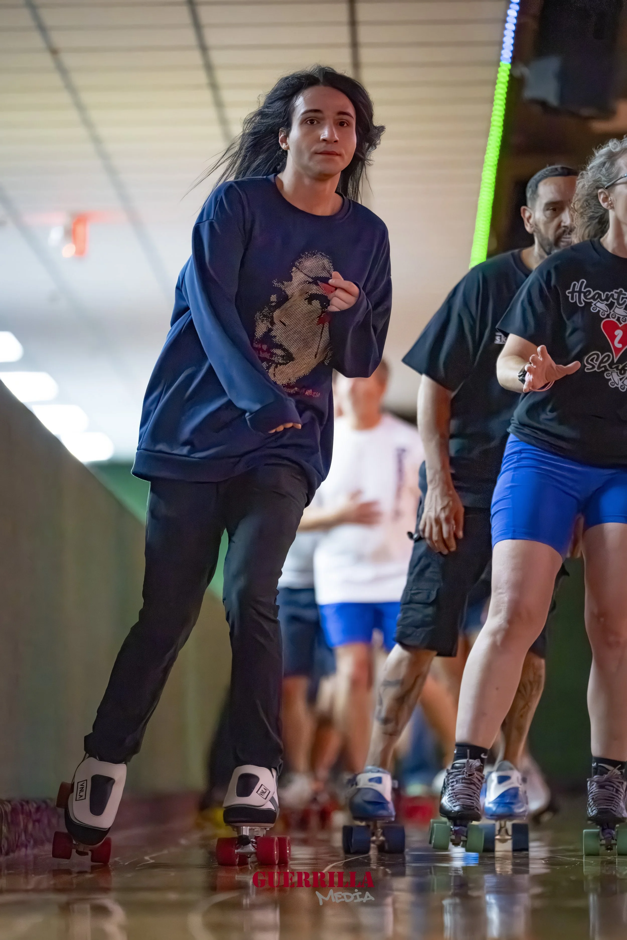 People roller skating indoors, with the main person being a woman wearing a black sweatshirt with a graphic print, black pants, and roller skates with white boots and pink wheels. Others in the background are skating as well.