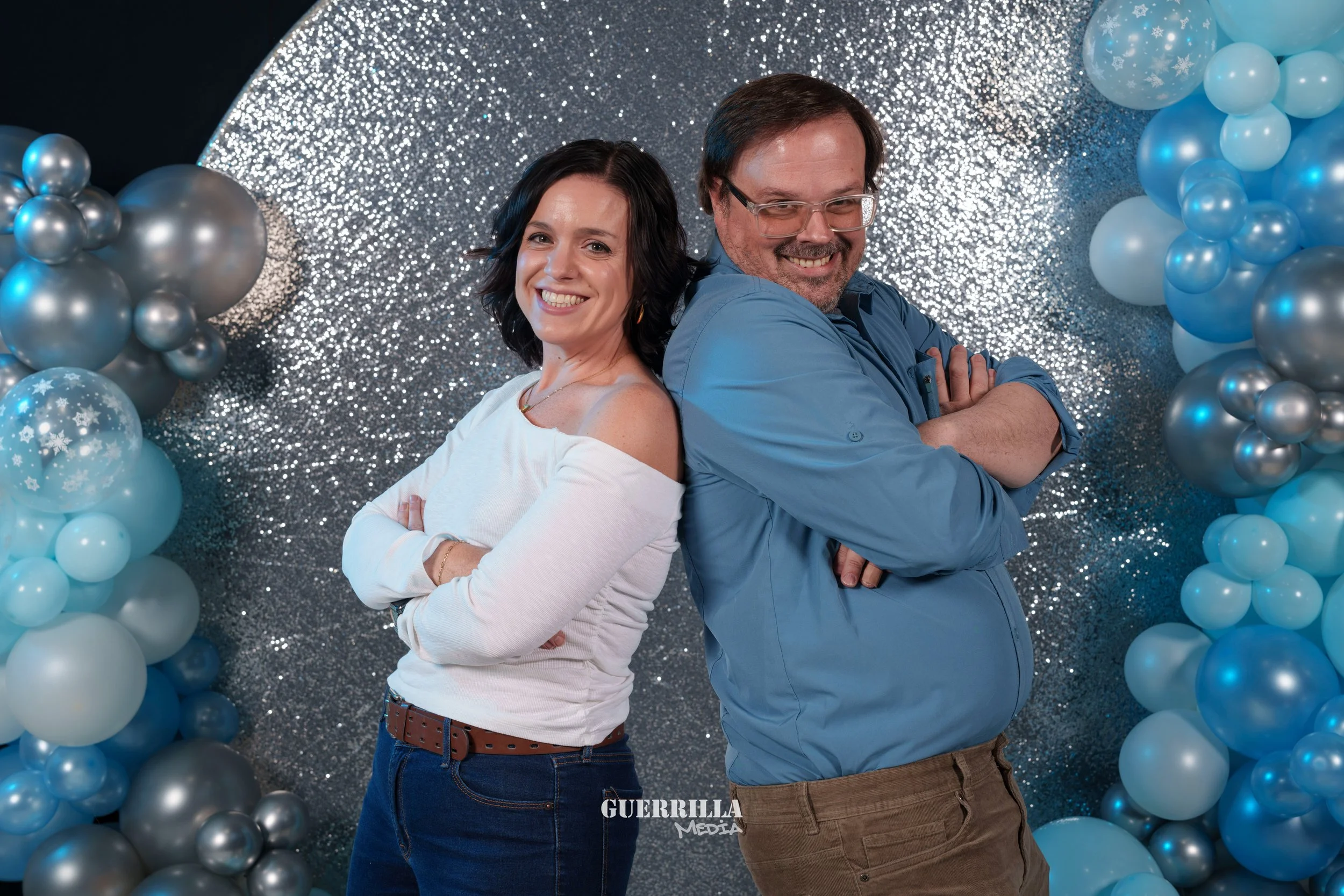A woman and a man standing back to back with arms crossed, smiling against a backdrop decorated with balloons and glittering silver backdrop.