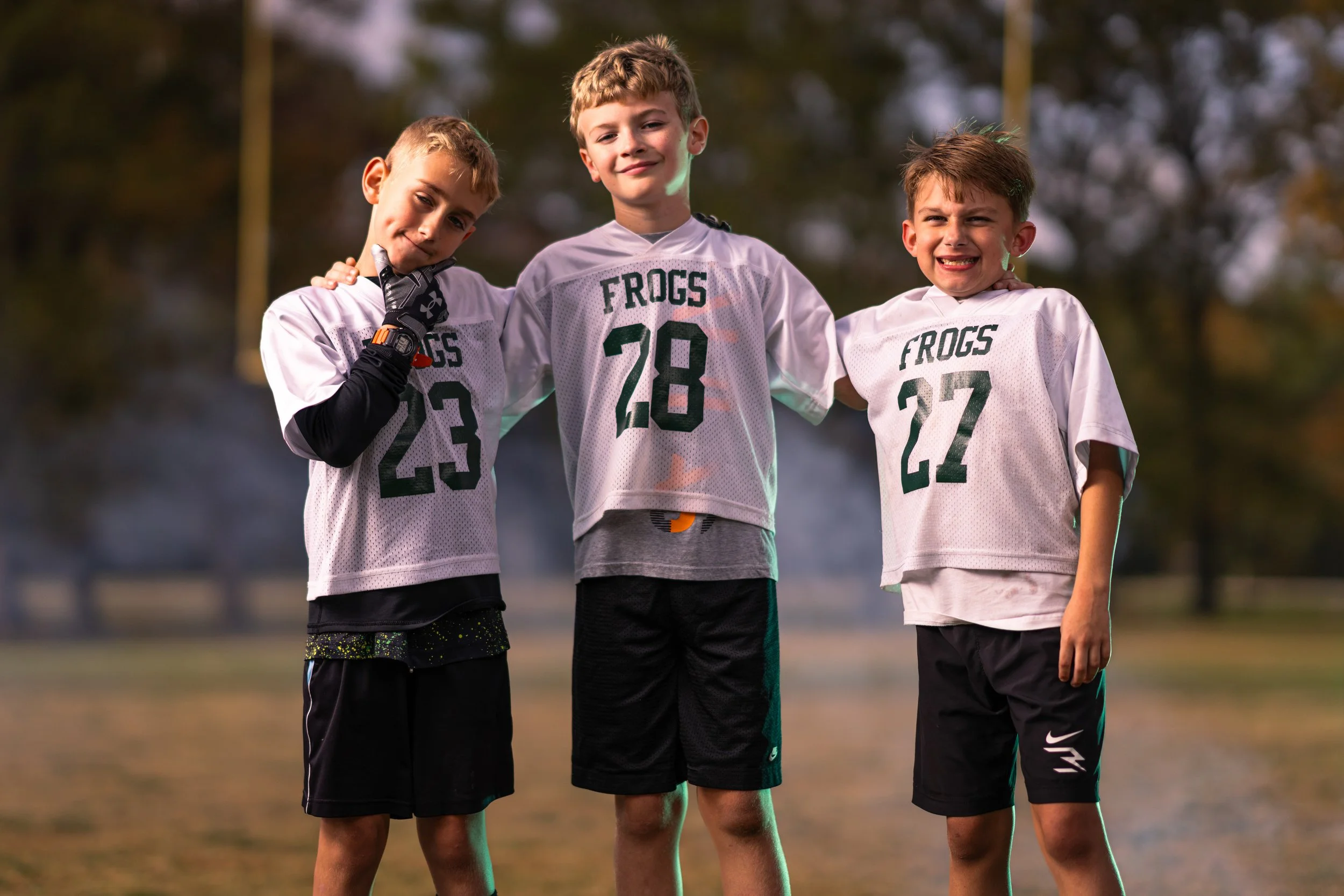 Three young boys in football uniforms from the FROGS team hugging each other on a football field during daytime.