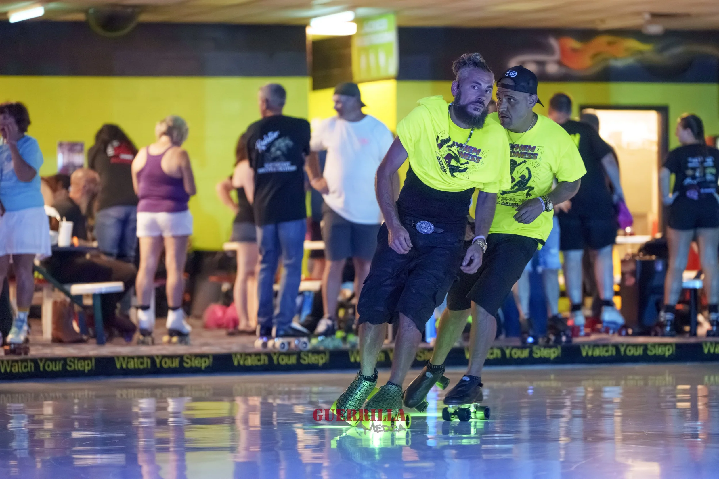 Two men skateboarding indoors at an event with a crowd in the background. They are wearing bright yellow shirts and are riding on glow-in-the-dark skateboards.