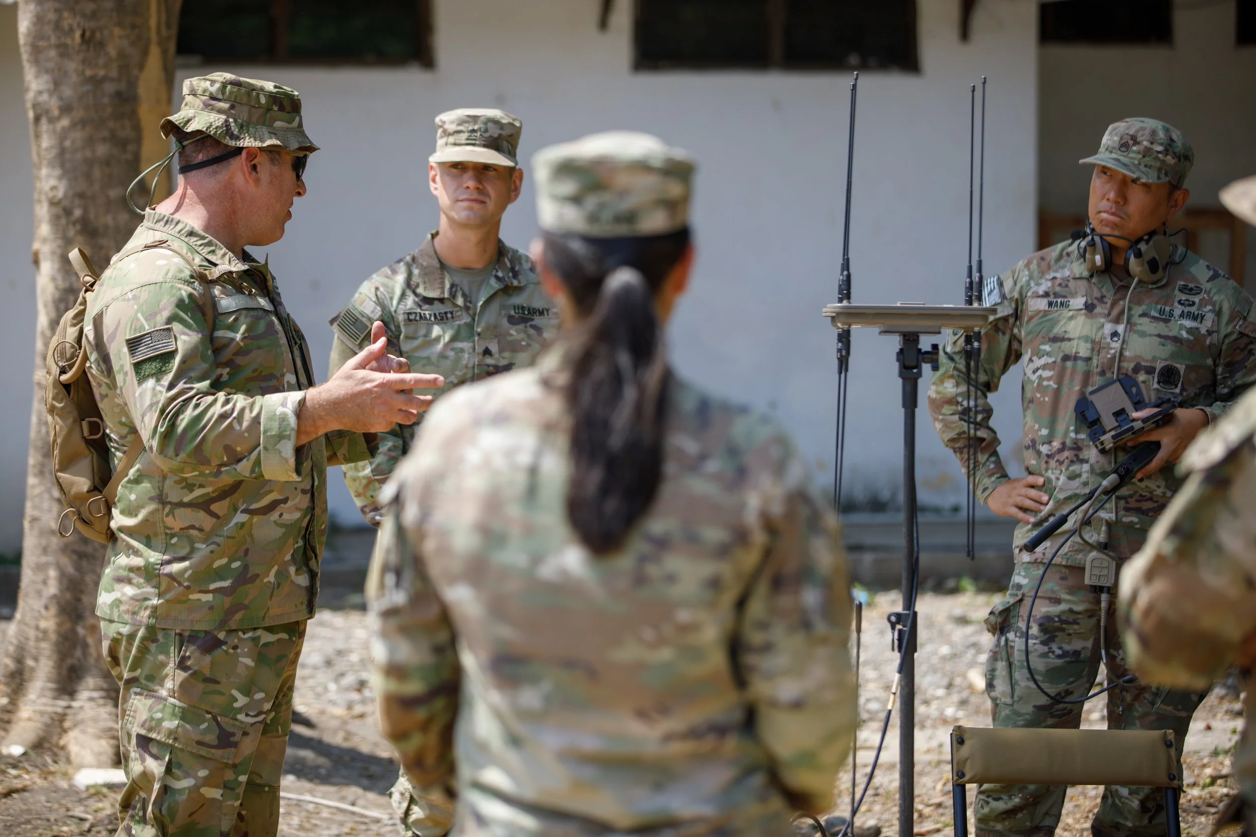 Group of military personnel in camouflage uniforms engaged in a discussion outdoors, with communication equipment and antennas present.