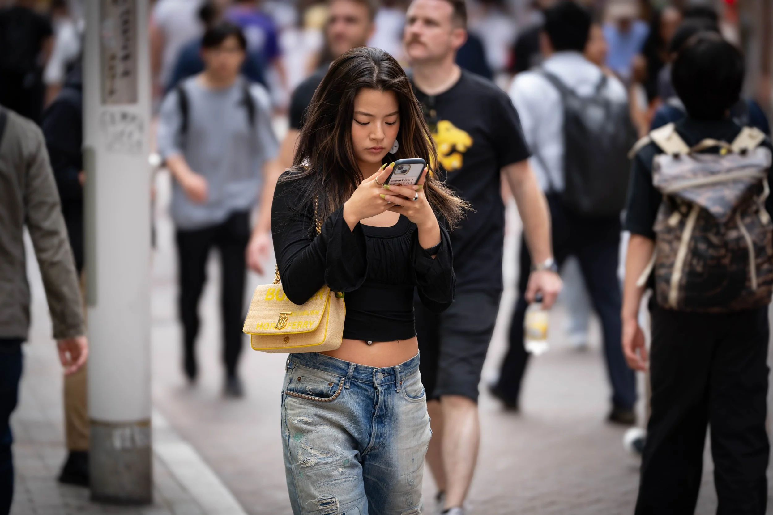 Young woman with long dark hair looking at her phone while walking on a busy city street surrounded by pedestrians.