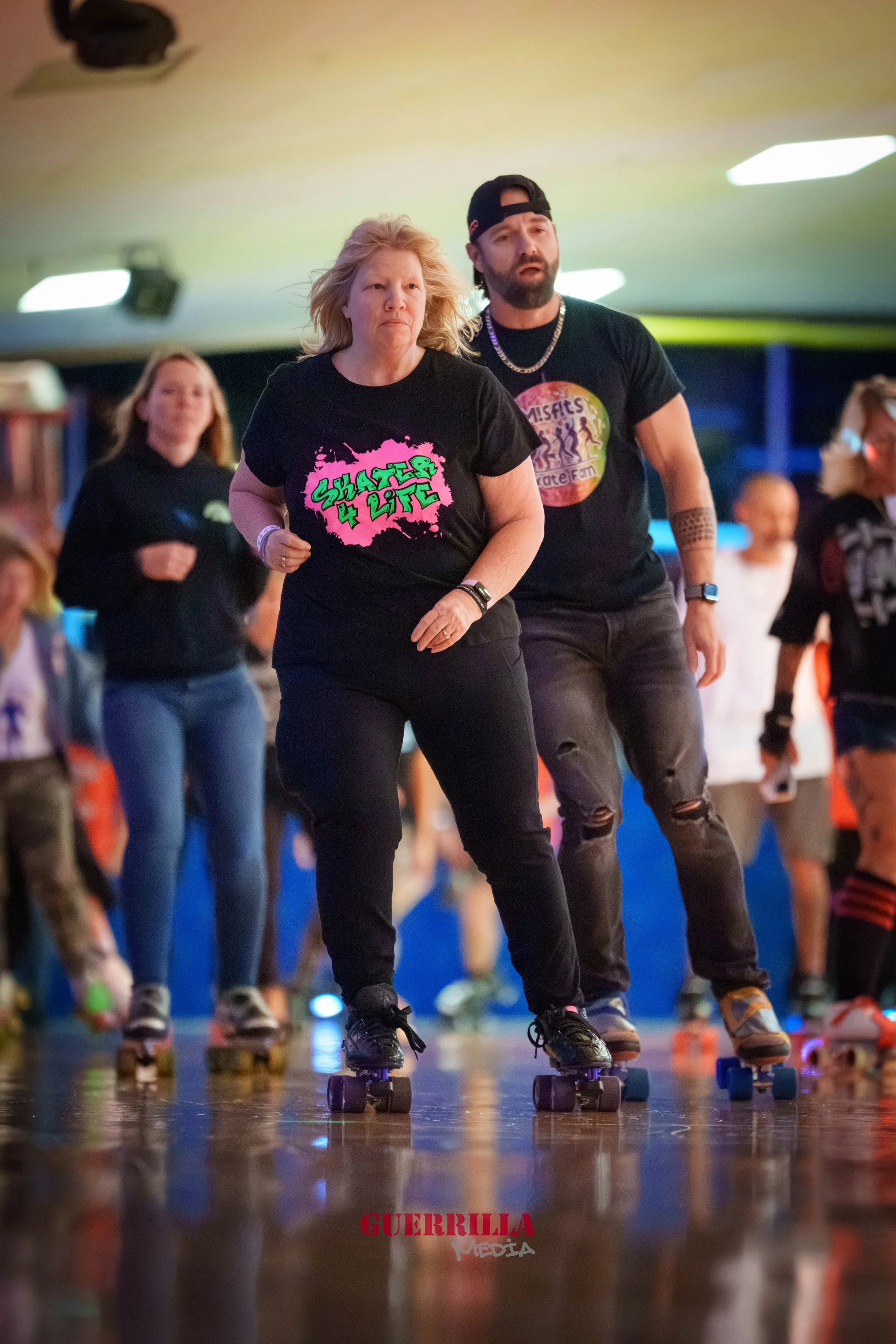 People roller skating in an indoor facility, with a woman in the foreground wearing a black t-shirt that says 'Skaters 4 Life' and a man behind her wearing a black t-shirt that says 'Misas Taste Farm.' Other skaters are visible in the background.