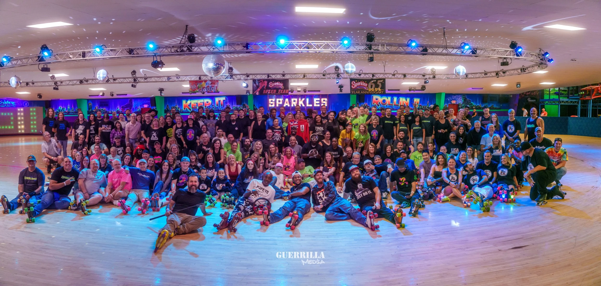 A large group of people gathered at a roller skating rink, posing for a group photo. The scene is vibrant with neon lights and signs in the background, including 'KEEP IT SPARKLES' and 'ROLLIN'. Many individuals are holding or wearing roller skates, 