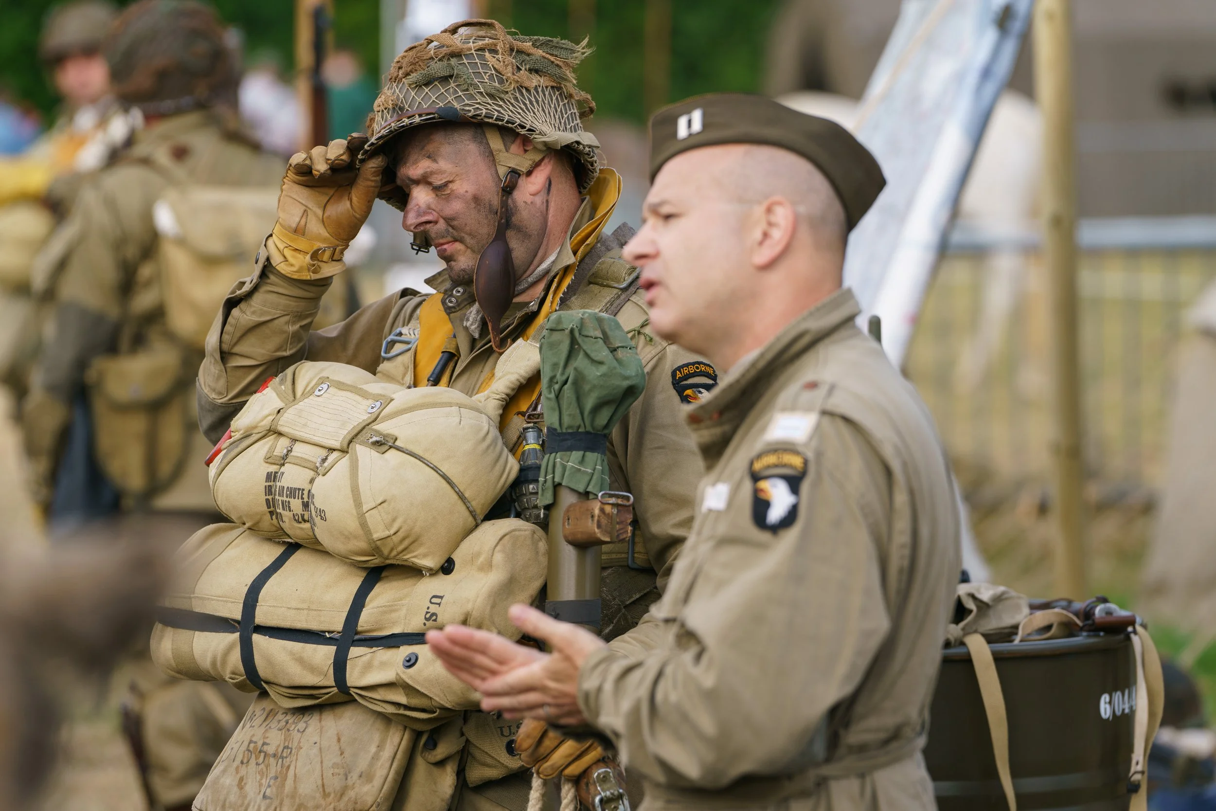 Two soldiers in military uniforms and gear, one with a helmet and the other with a cap, are engaged in a moment of prayer or reflection outdoors. There are other soldiers in similar uniforms in the background.