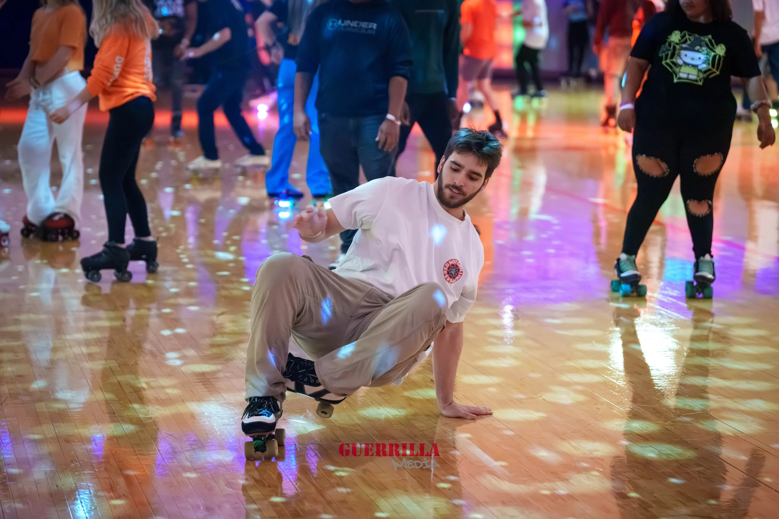 A man roller skating on a wooden floor at an indoor skating rink. He is leaning to one side with one hand on the floor and the other in the air, smiling. Several other people are roller skating in the background with colorful lights reflecting on the