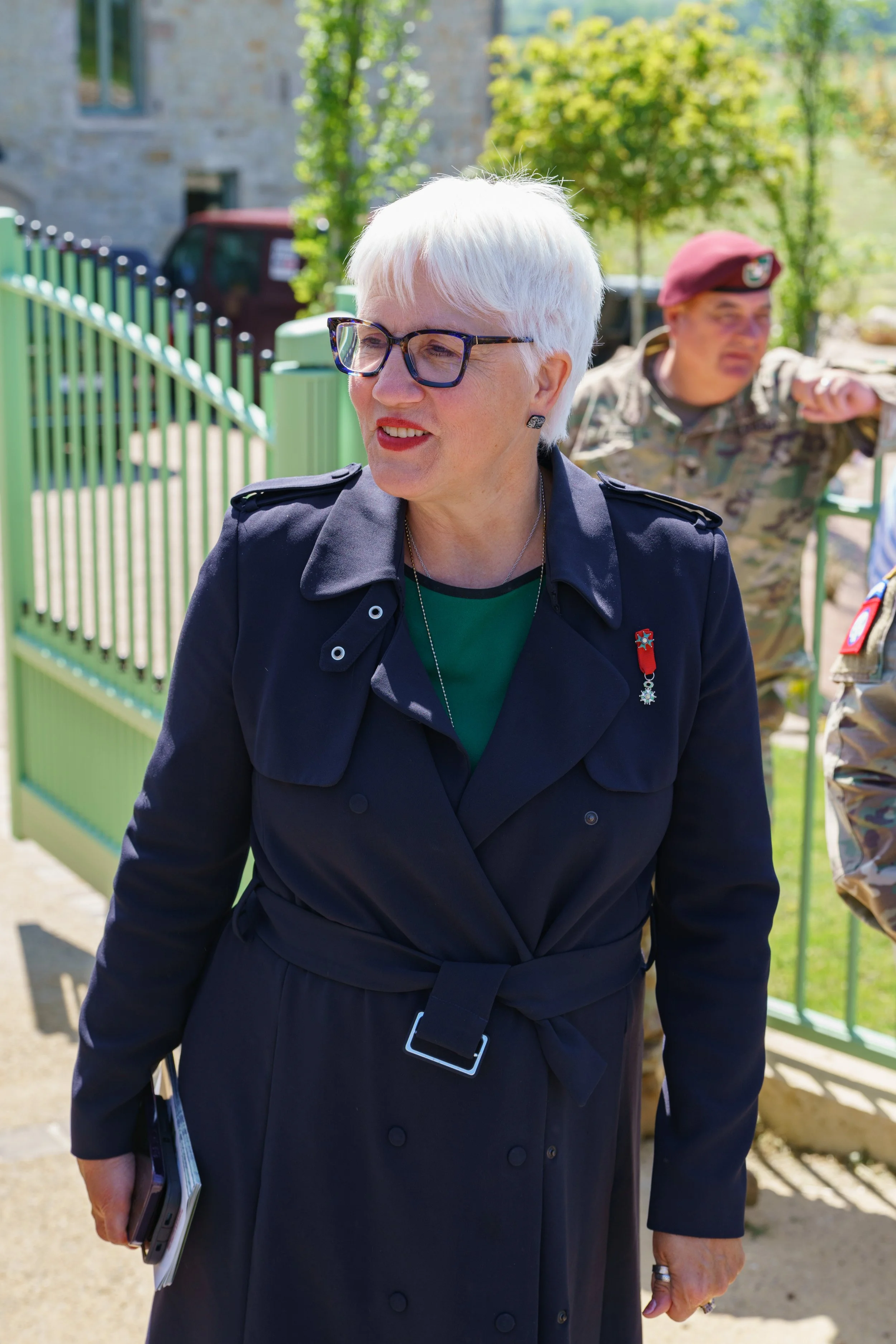 An older woman with short white hair wearing glasses and a dark coat with a medal pin, standing outdoors on a sunny day. In the background, a man in camouflage military uniform with a maroon beret is leaning against a fence.