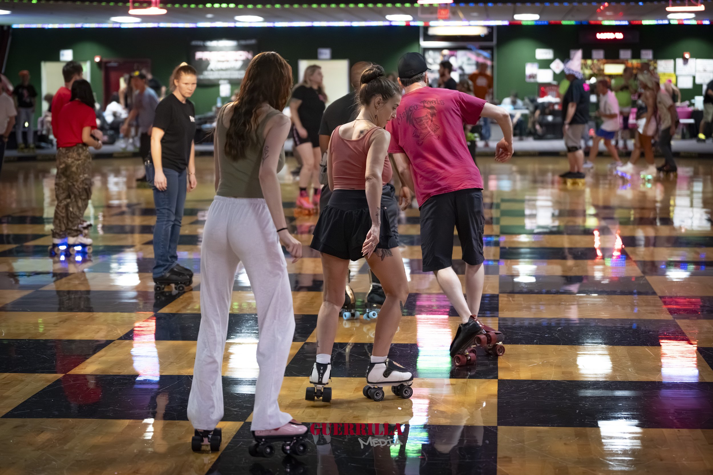 People roller skating indoors on a polished, checkered floor with colorful lights and a neon sign in the background.