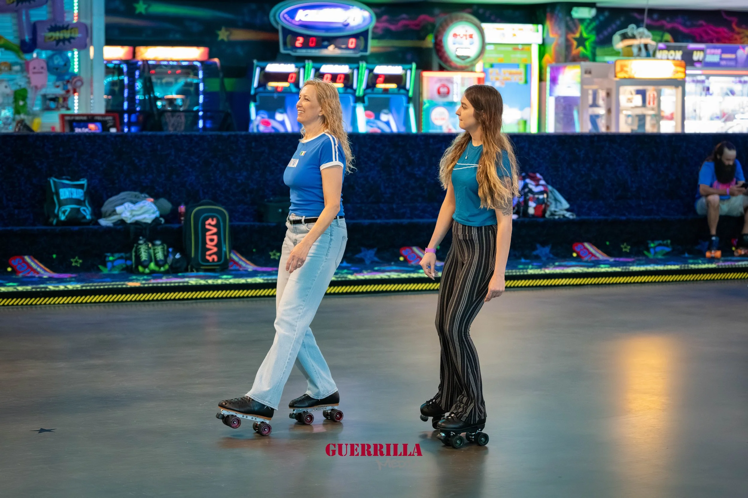 Two women roller skating in an indoor arcade or roller rink, colorful neon lights, arcade games in the background, one with blonde hair and light blue jeans, the other with long brown hair and striped pants.