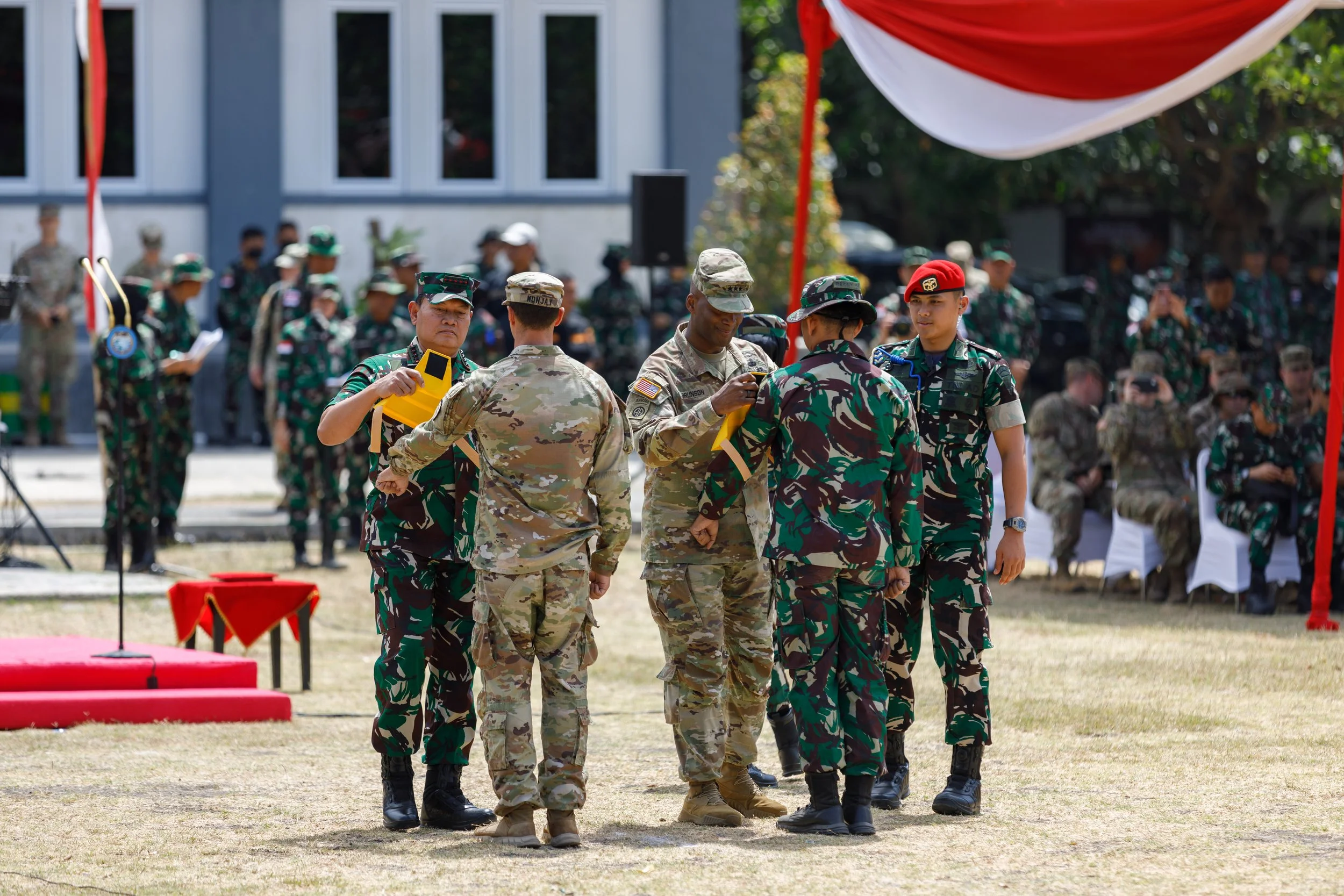 Military personnel in camouflage uniforms during a ceremonial event, with soldiers standing in the background and a red and white draped banner overhead.