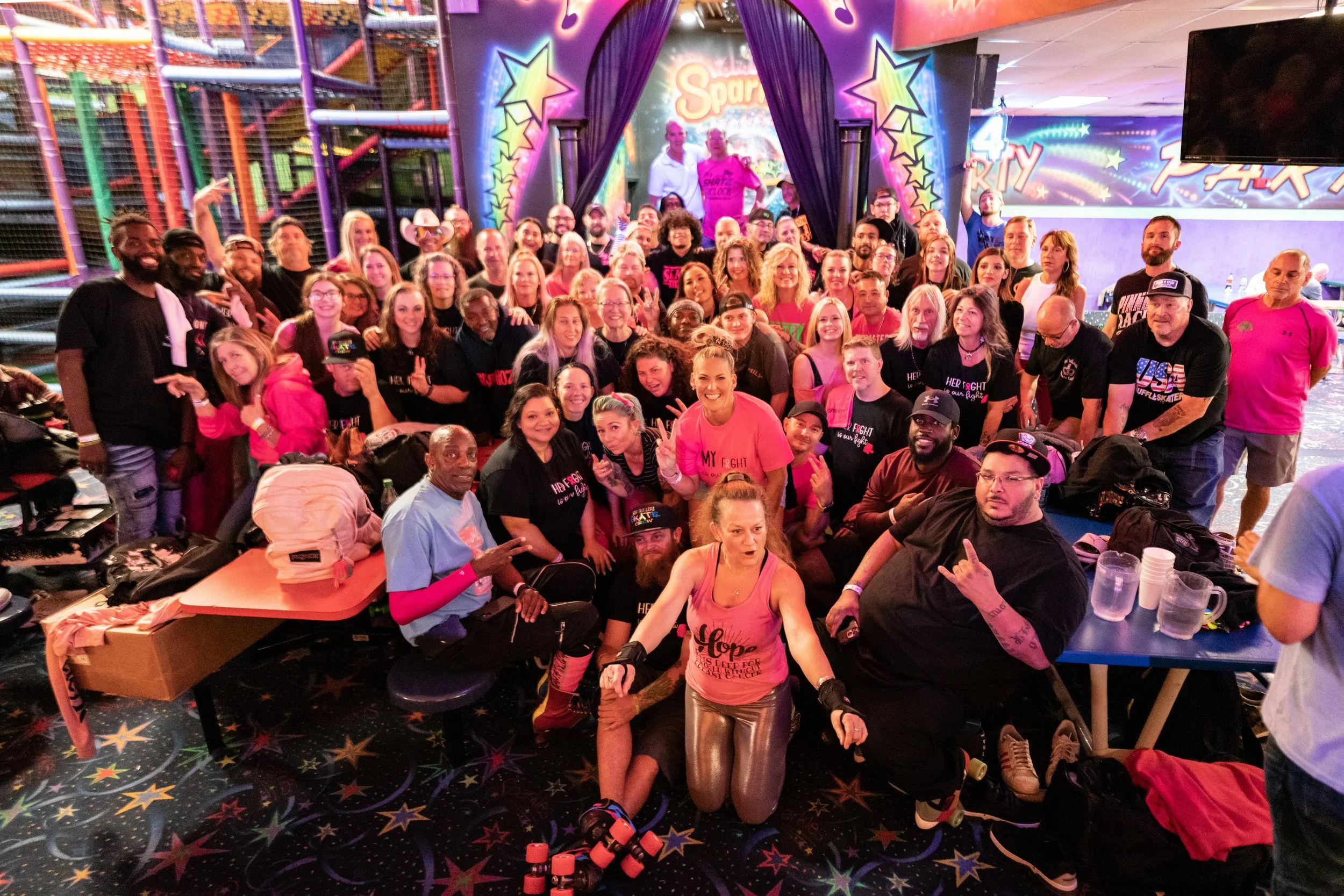 A large group of people gathered inside an arcade or entertainment center with colorful lights and decorations, posing for a group photo. The background features a stage with neon stars and the word 'Spark.' Some people are wearing matching pink shir
