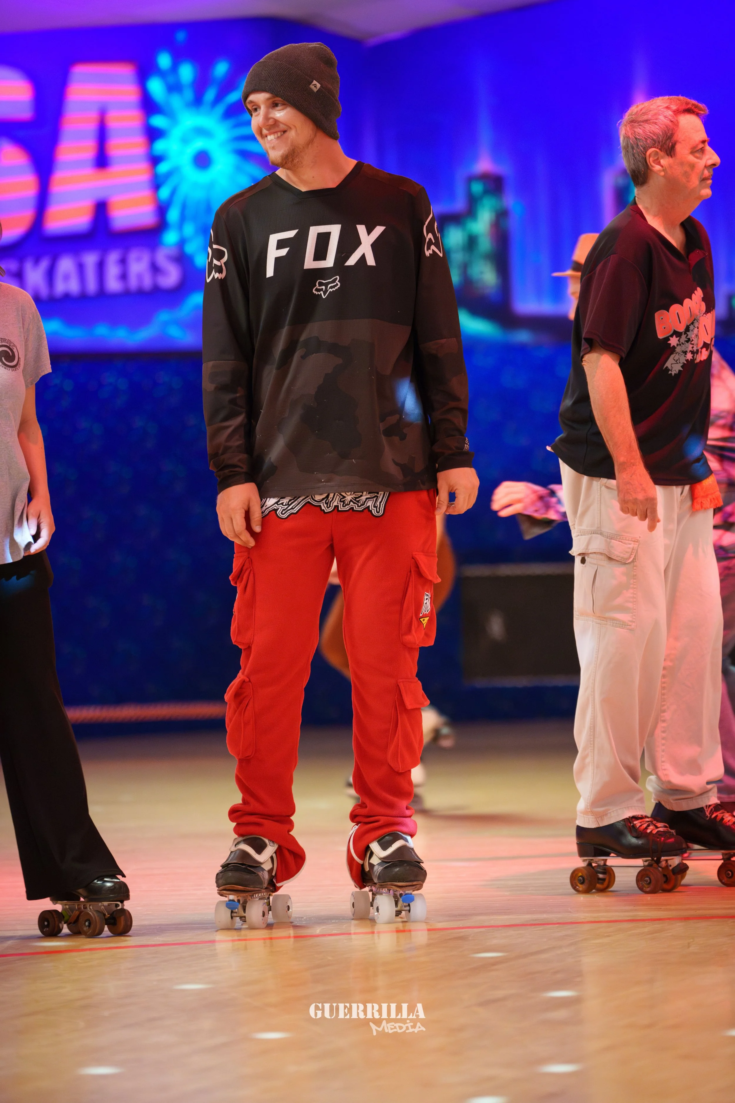 A group of people roller skating indoors, with a blue and purple backdrop featuring a firework display and the words 'SKATERS'. The focus is on a smiling man wearing a gray beanie, black and camo sweatshirt with 'FOX' in white letters, and bright red