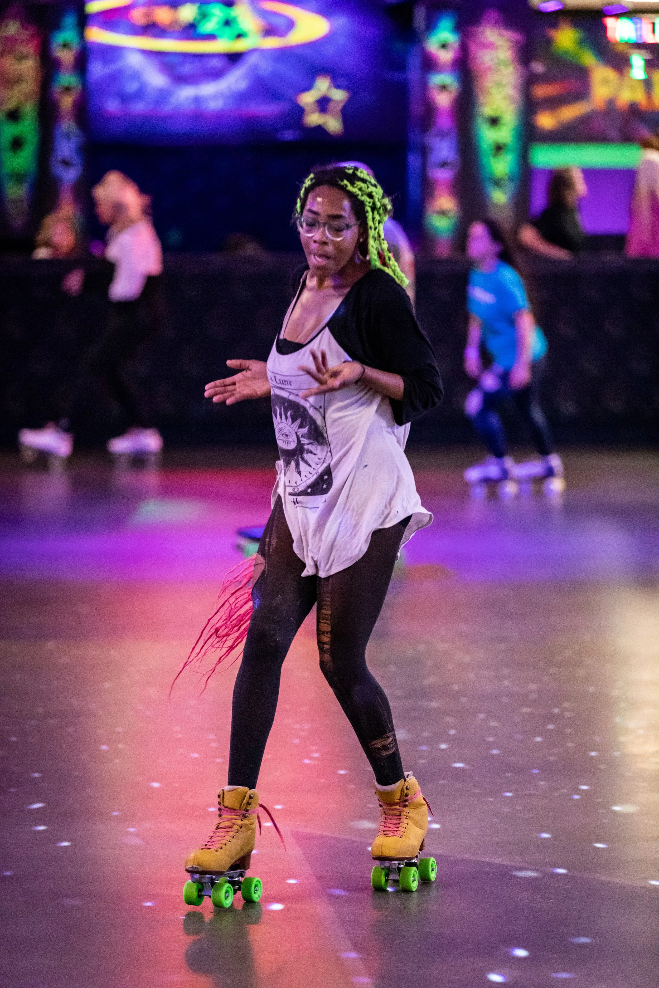 A woman roller skating wearing yellow shoes, black leggings, a white oversized T-shirt with a graphic print, and a black long-sleeve shirt underneath at an indoor roller skating rink with colorful neon lights.