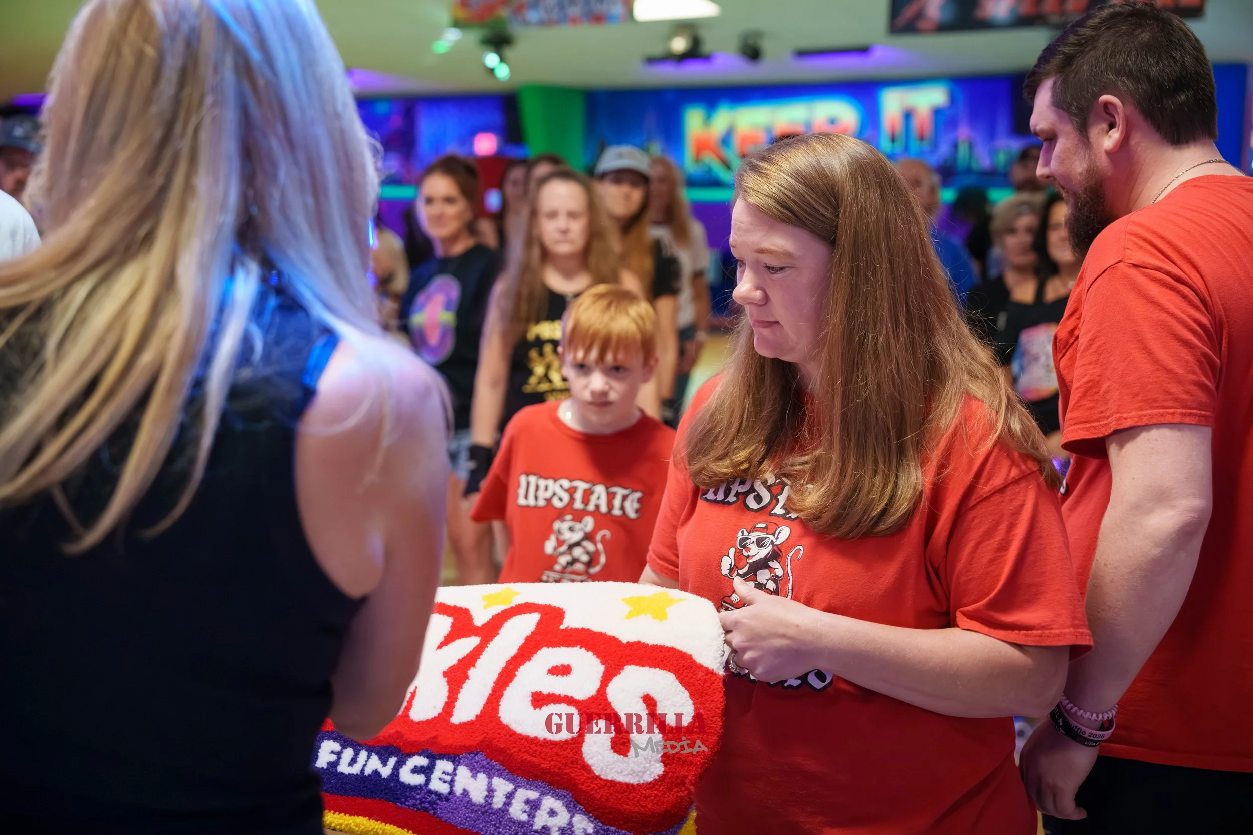 A group of people gathered at an arcade or entertainment center, with a woman holding a large red and white woven blanket or banner. The background features colorful neon signs and a crowd observing the scene.