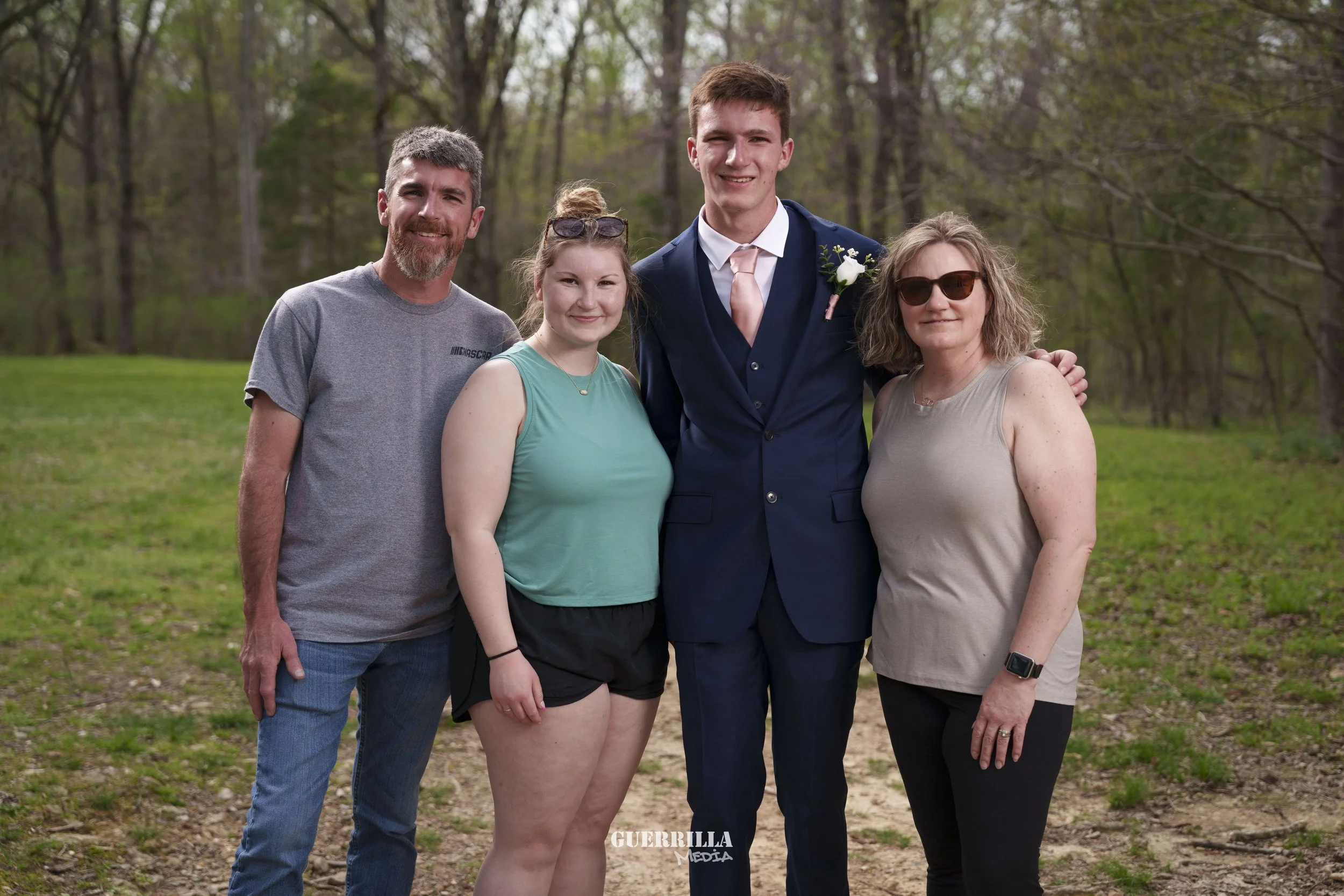 A group of five people standing outdoors on a grassy path in a wooded area. The group includes two men and three women, with the man in the center wearing a navy blue suit with a pink tie, suggesting a formal occasion like a wedding. The others are d