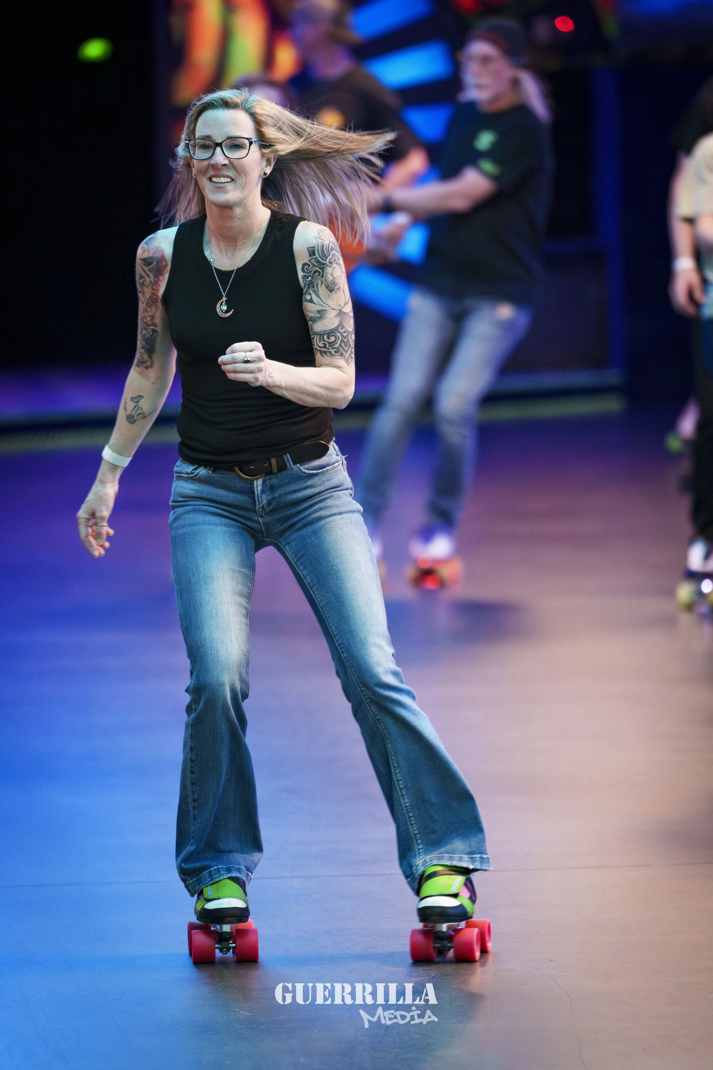 Woman with long hair, glasses, and tattoos roller skating indoors with friends, brightly lit with colorful background lights.