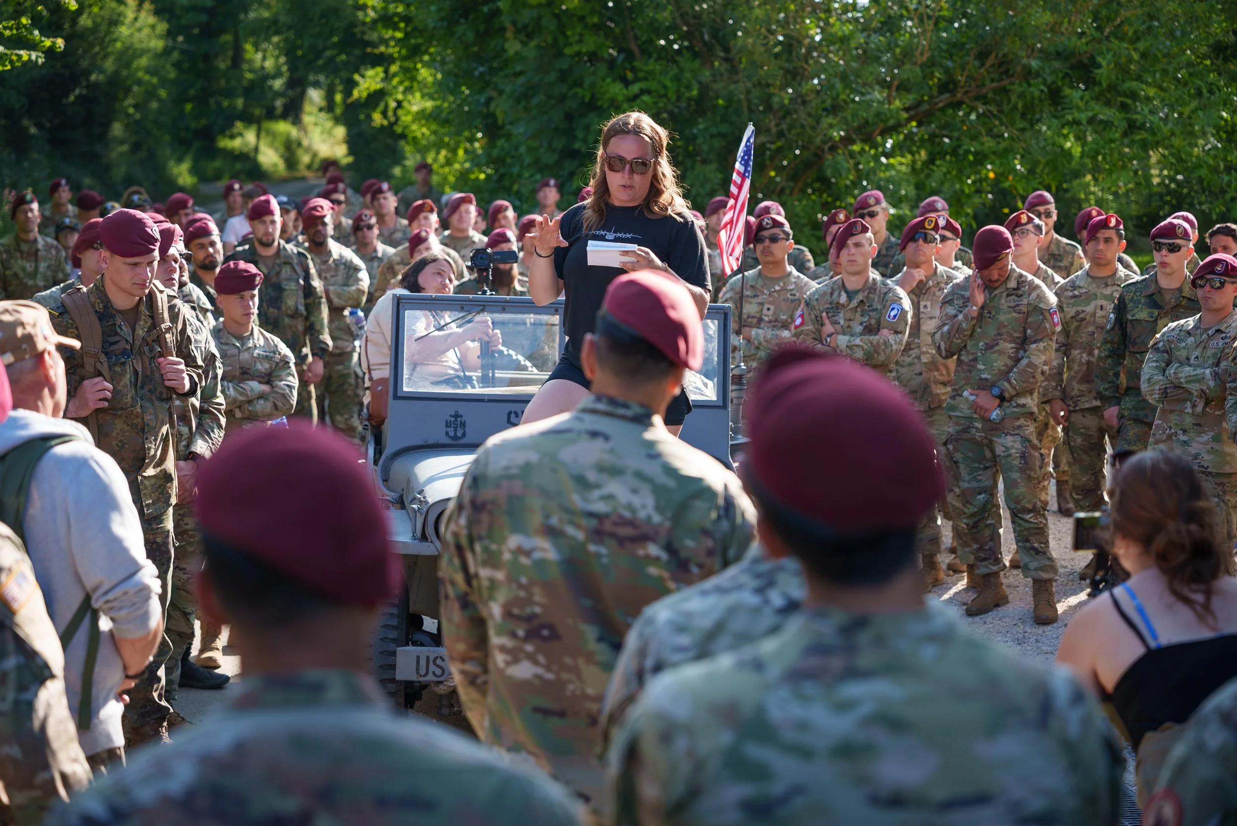 A woman is speaking to a crowd of soldiers wearing camouflage uniforms and maroon berets, with some soldiers saluting and others listening, outdoors with trees in the background.
