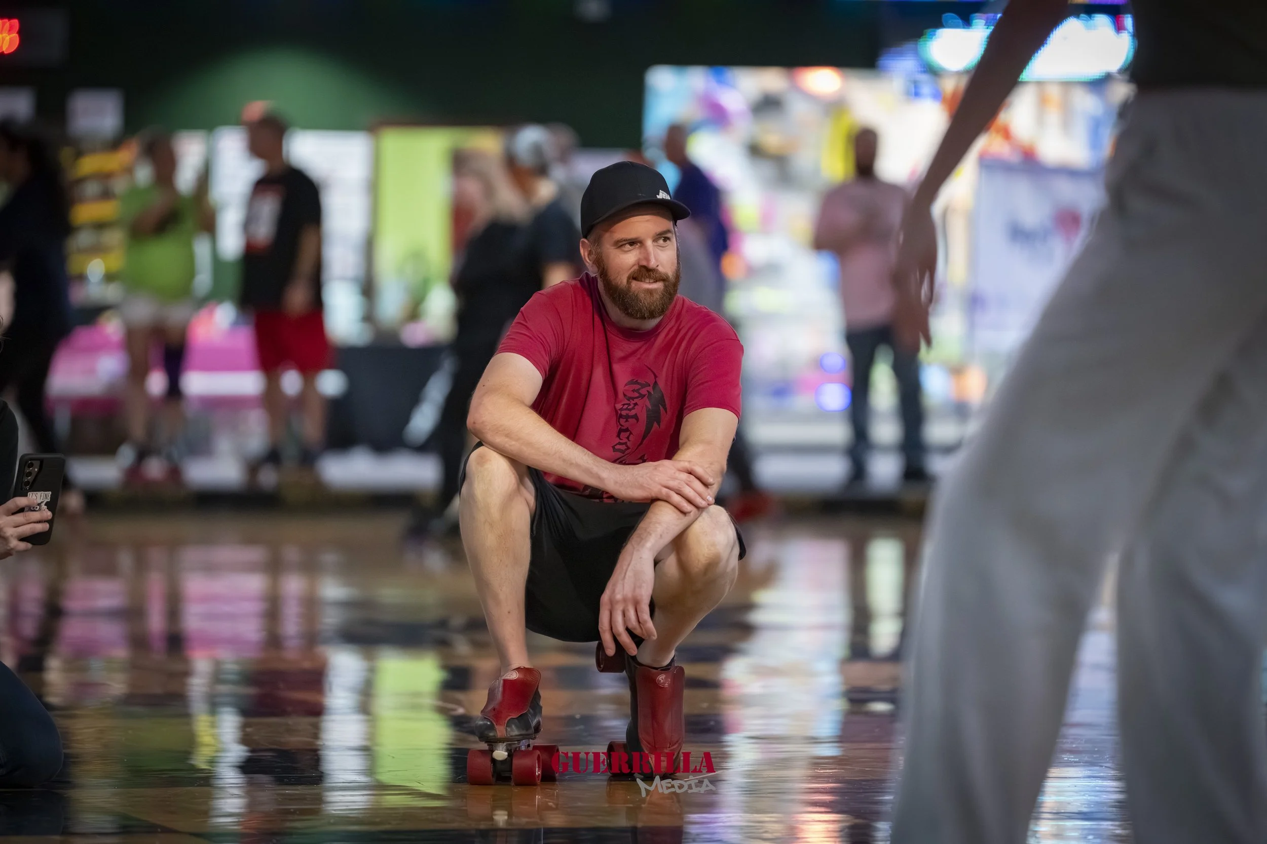 A man in a red t-shirt and black shorts crouches on a skateboard inside a roller-skating rink, with people and colorful displays blurred in the background.