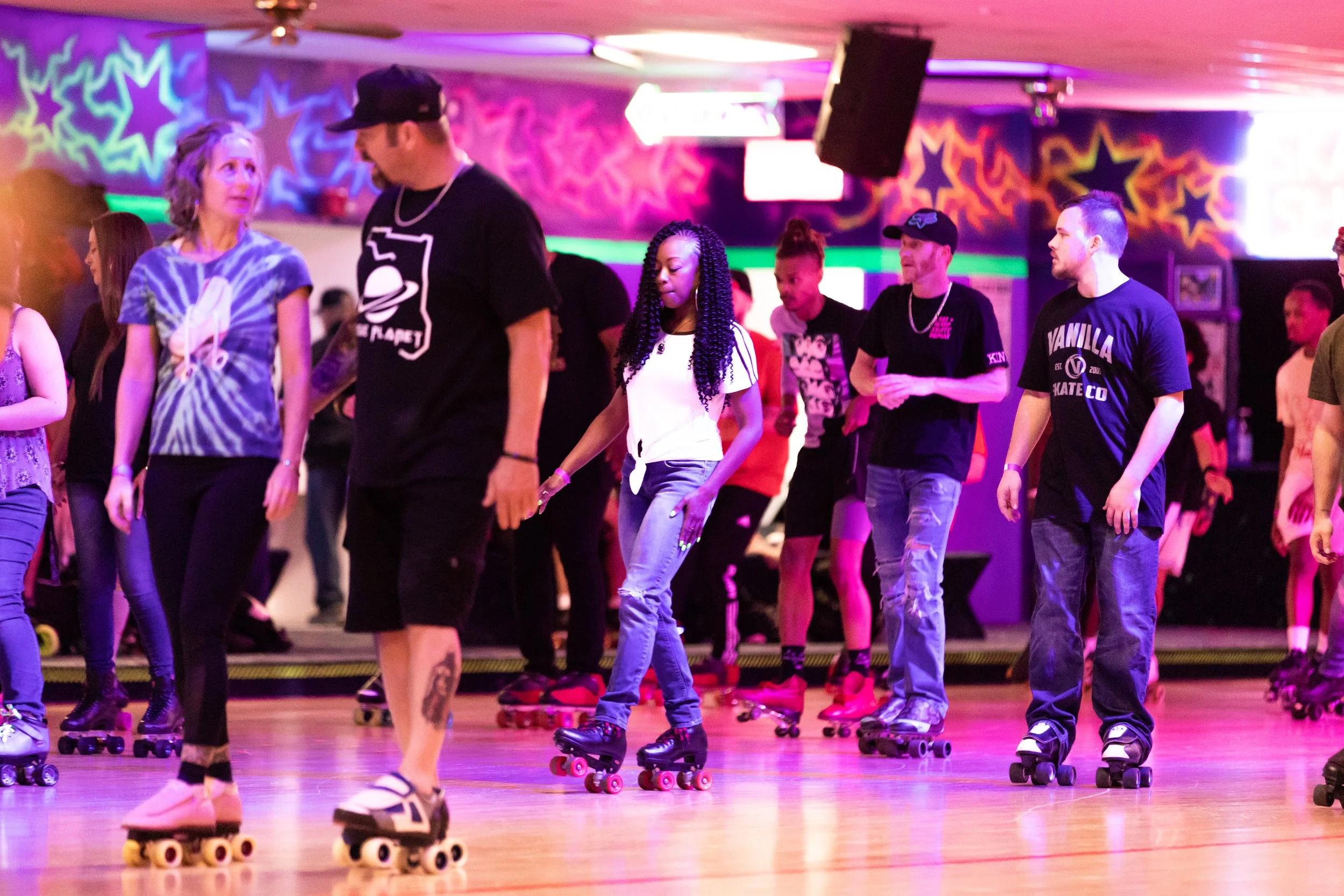 People roller skating at an indoor roller rink with colorful star and swirl wall art, neon signs, and soft pink and purple lighting.
