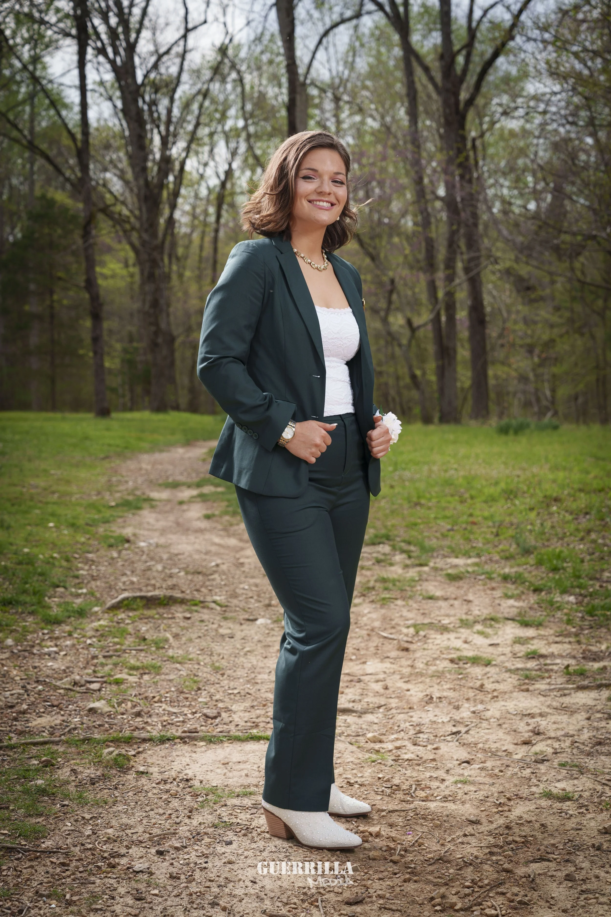 A woman in a dark green business suit with a white top, standing on a dirt path in a forest with green trees and a cloudy sky behind her, smiling with hands in pockets.