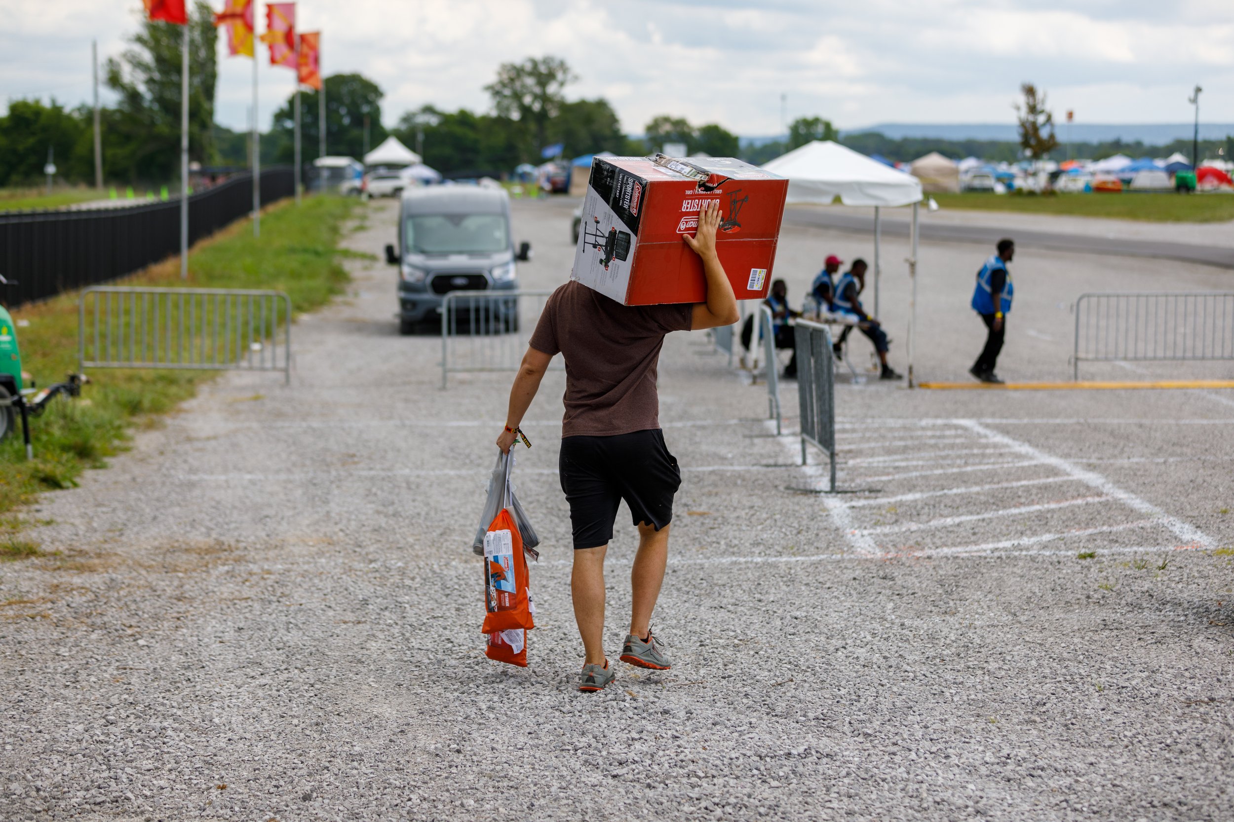 Person carrying a large boxed grill on his shoulder and a smaller item in his hand at an outdoor event with tents, flags, and several people in the background.