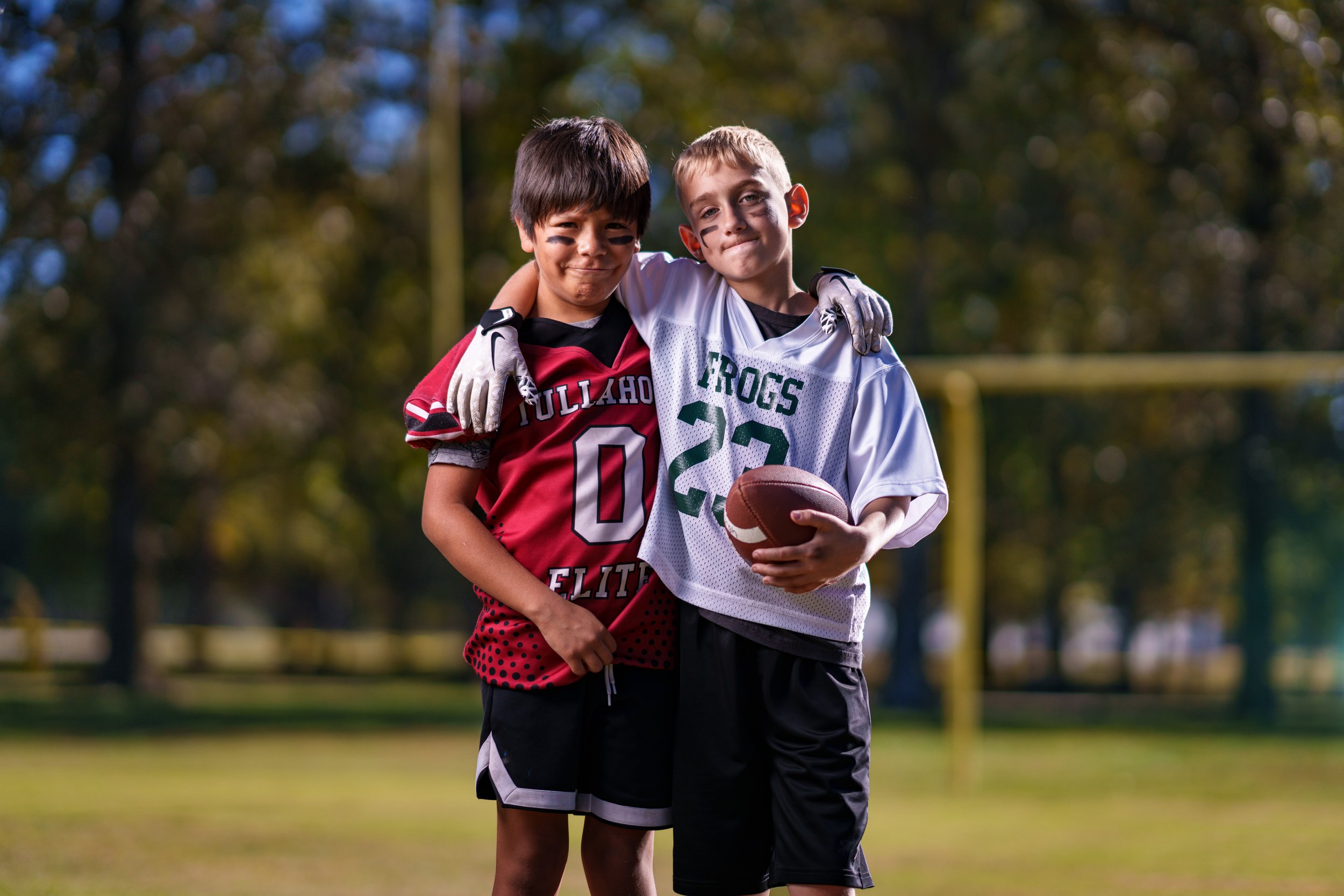 Two young boys stand close together on a football field, smiling with their arms around each other's shoulders. One boy wears a red football jersey with black and white accents, and the other wears a white football jersey with green lettering and the
