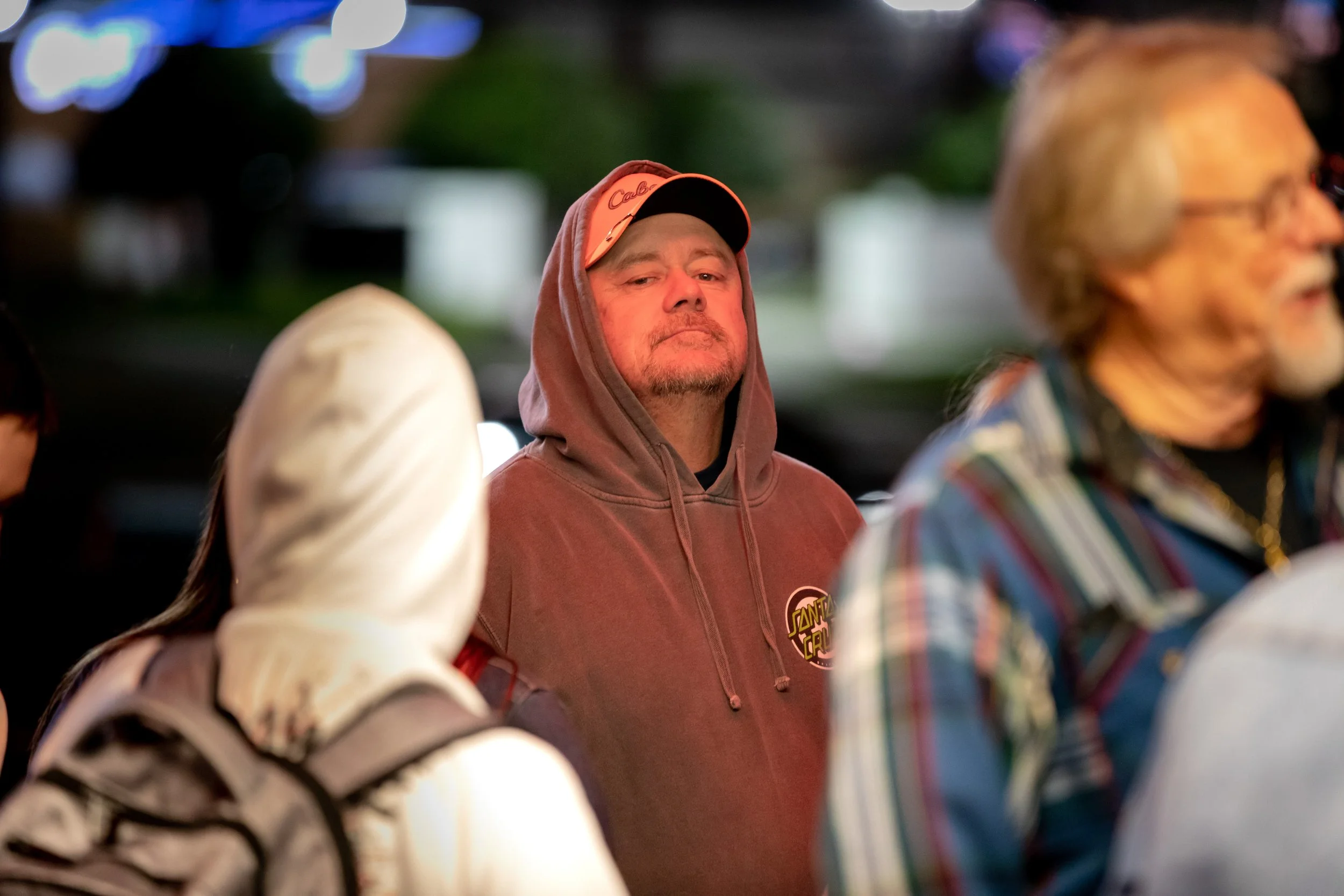 A man in a maroon hoodie with a Santa Cruz logo and a cap underneath, standing among a group of people at night.