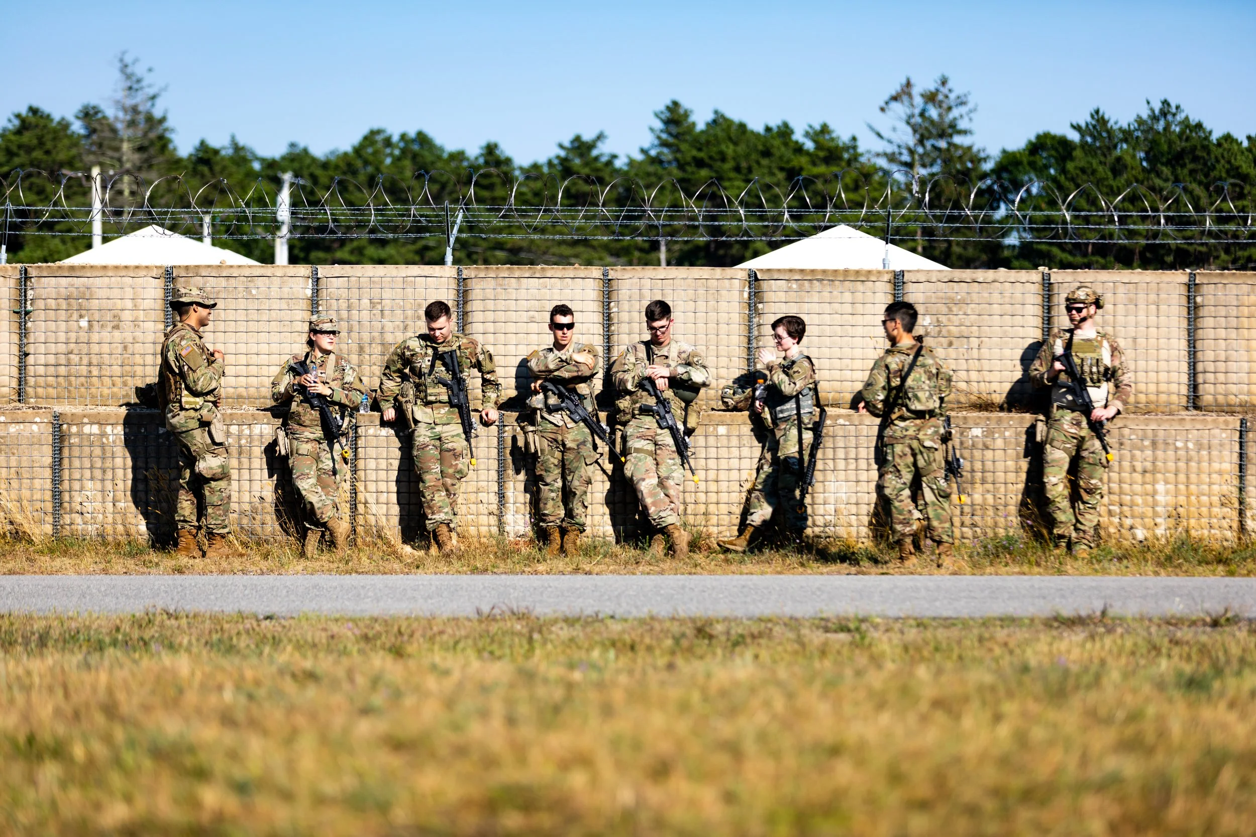 Group of soldiers standing along a wire-topped fence outdoors on a sunny day