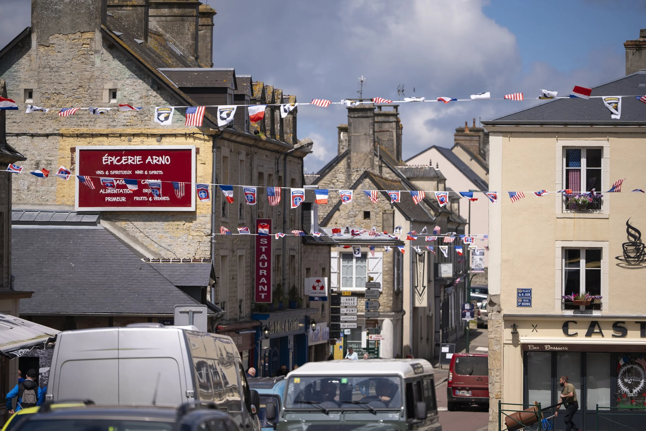 A street scene with French flags and patriotic bunting hanging across the road in a small town, with cars parked and a person pushing a cart.
