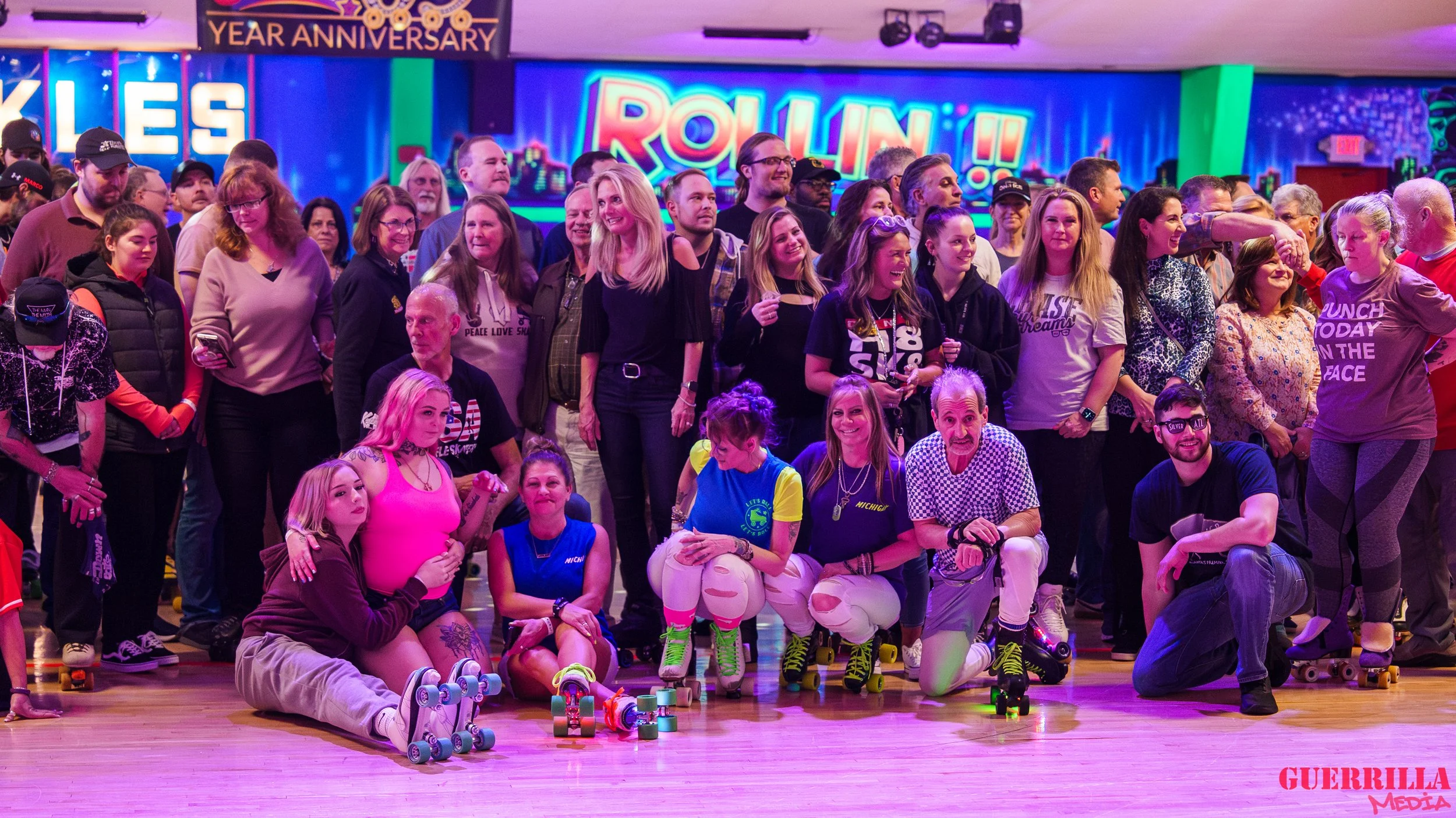 A large group of people celebrating at a roller skating rink, with some sitting on the floor and others standing behind them. Some are wearing roller skates, and there are colorful neon signs in the background, including one that says 'ROLLIN!!' and 
