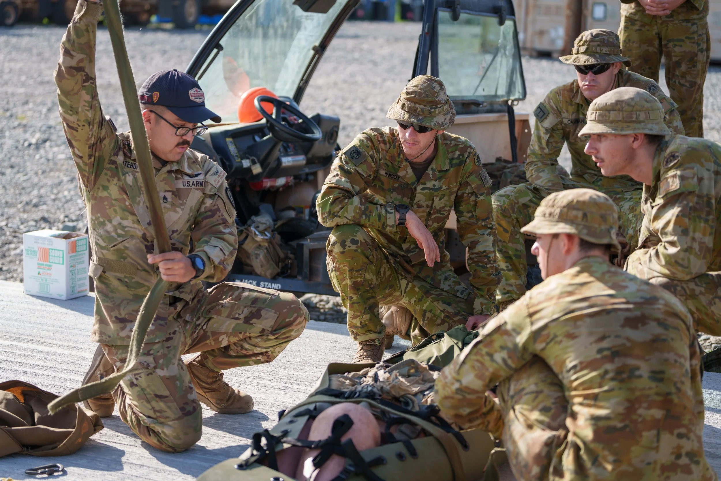 Group of soldiers in camouflage uniforms gathered outdoors, sitting or kneeling on a concrete surface, with military equipment and a small vehicle nearby, engaged in discussion or training.