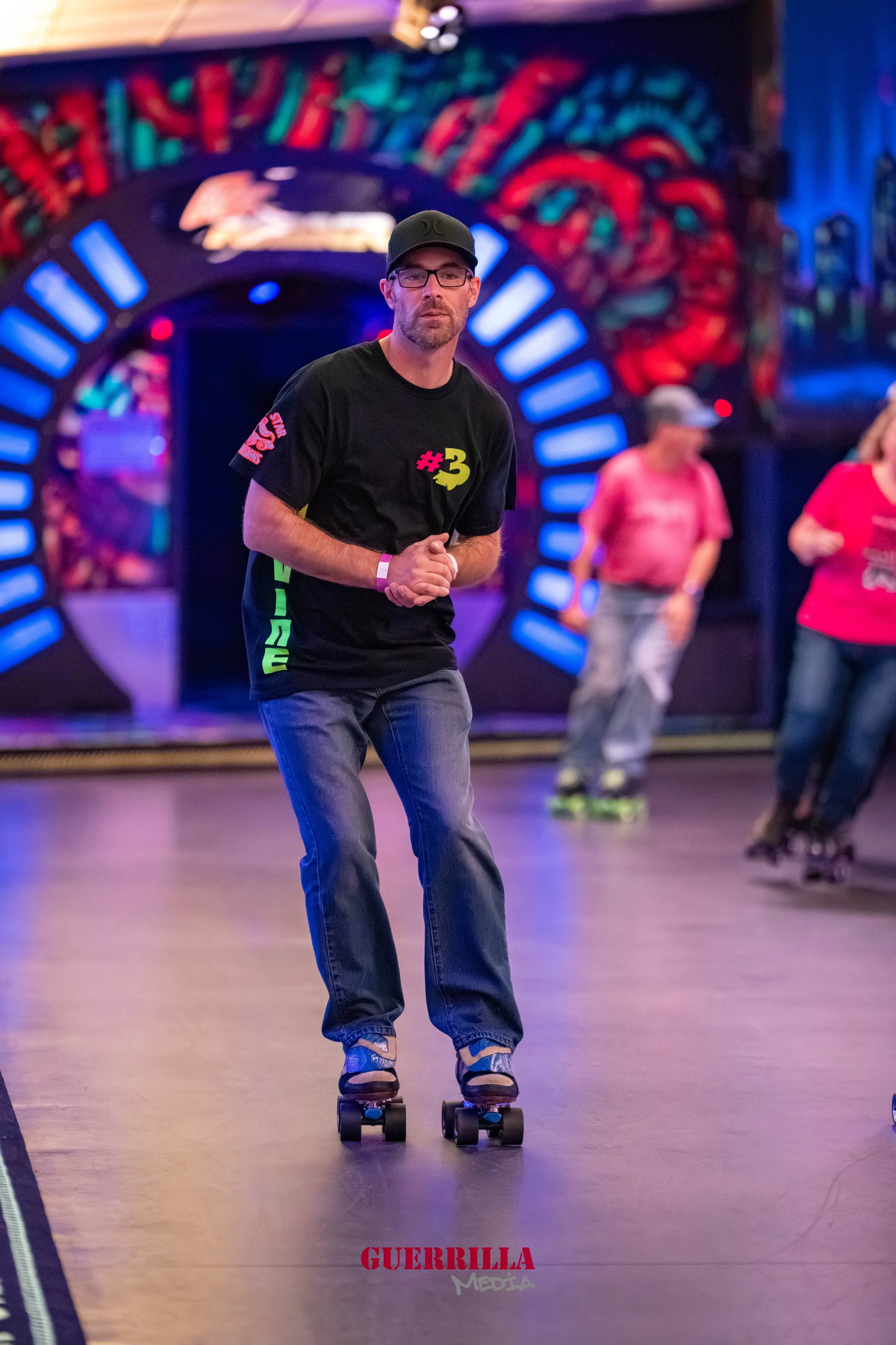 A man riding a skateboard in an indoor roller skating rink with colorful graffiti and lights in the background. He is wearing a black cap, glasses, a black t-shirt with colorful text, and jeans.