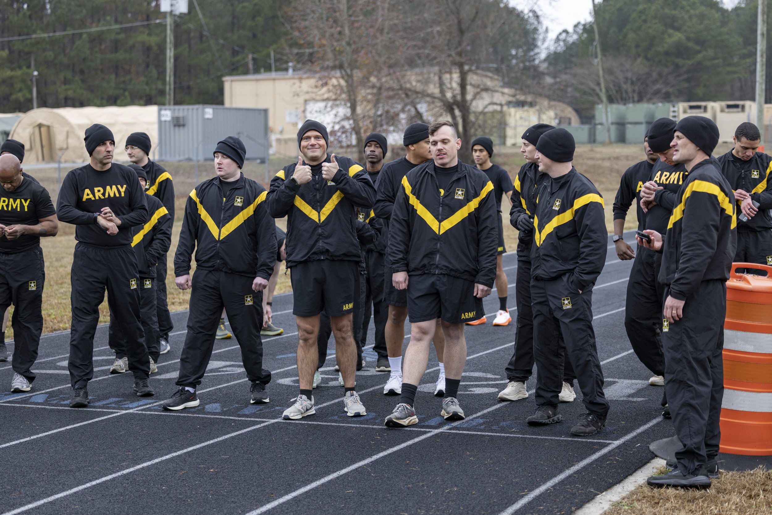 A group of male athletes wearing black and yellow army jackets and black beanies, standing on a running track, some preparing for a race, with trees and equipment in the background.