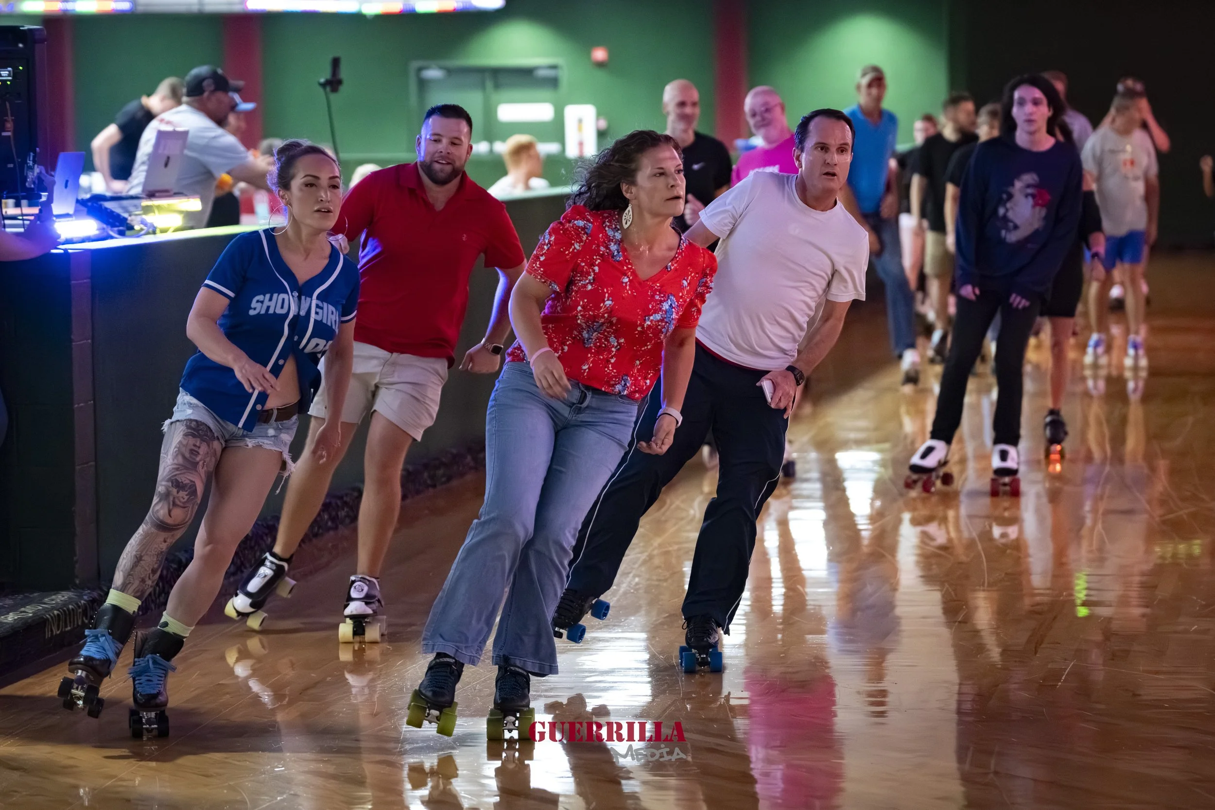 People roller skating inside a roller rink with a green backdrop and wooden floor, some wearing casual clothes, and a few in sportswear, with one woman wearing a blue jersey and denim shorts, and a woman with a red floral shirt.