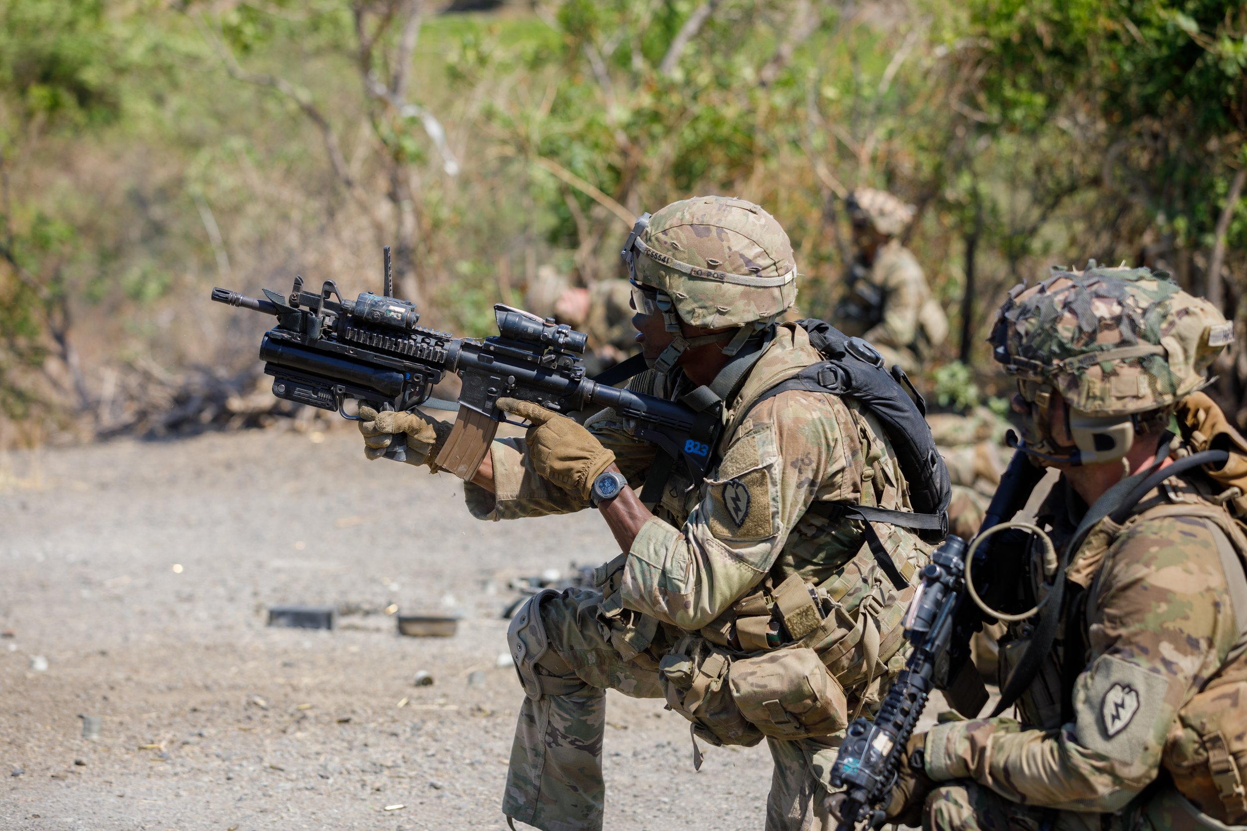 Three soldiers in camouflage gear during a training exercise, with two aiming rifles and one in the background in a wooded outdoor setting.