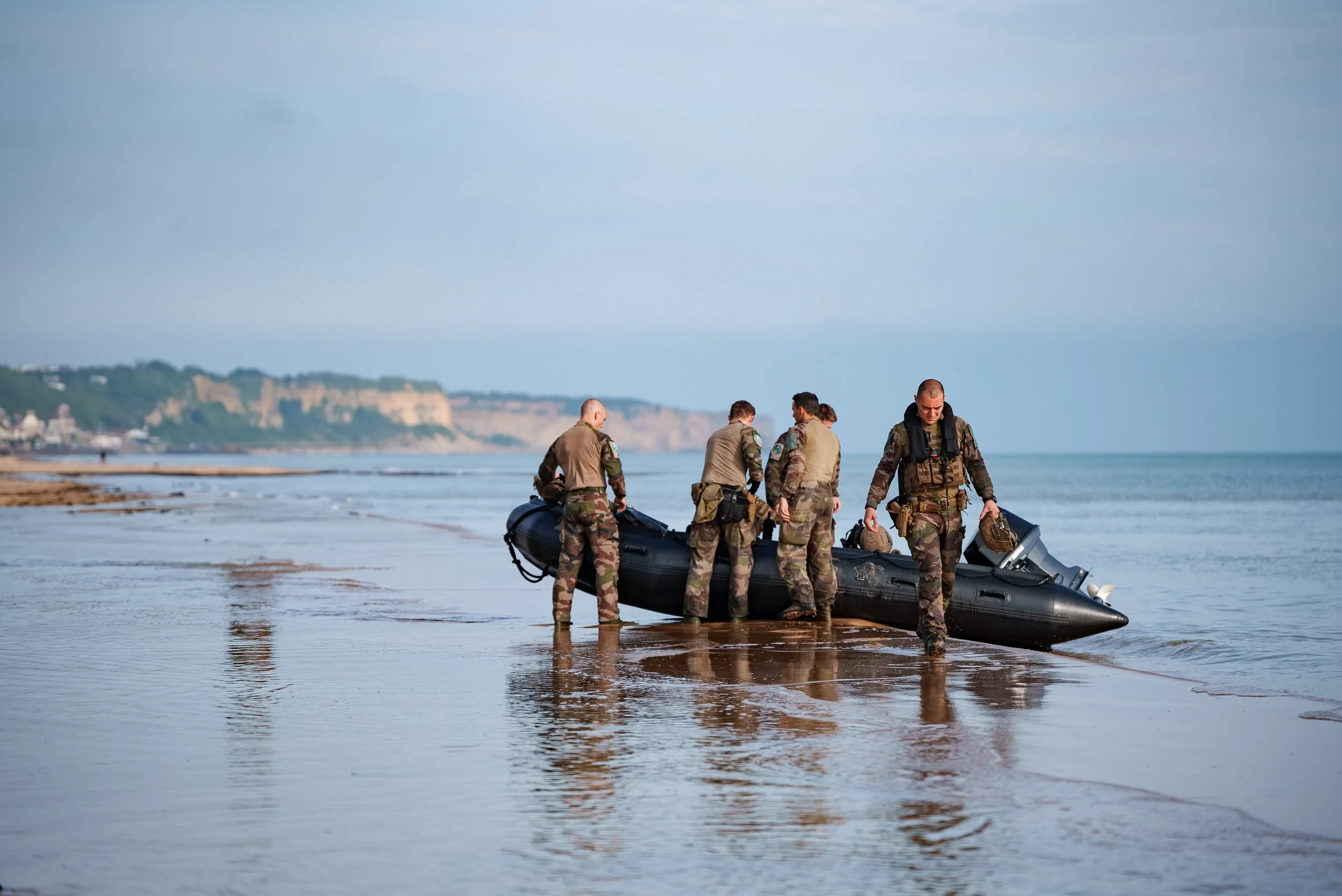 Military personnel in uniform launching a black inflatable boat into the ocean near the shore.