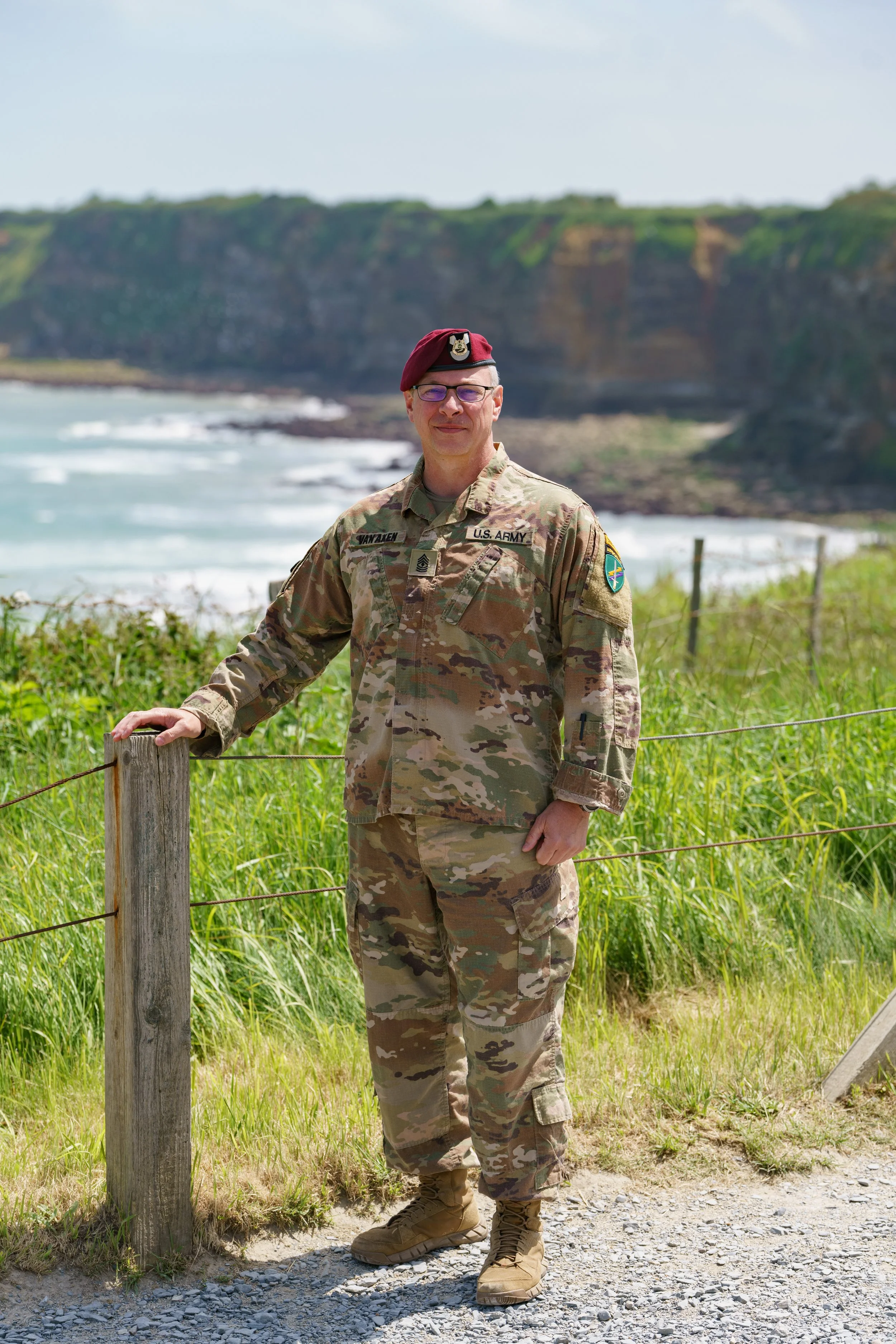 Man in U.S. Army camouflage uniform and maroon beret standing outdoors near a grassy fence with a coastal landscape in the background.