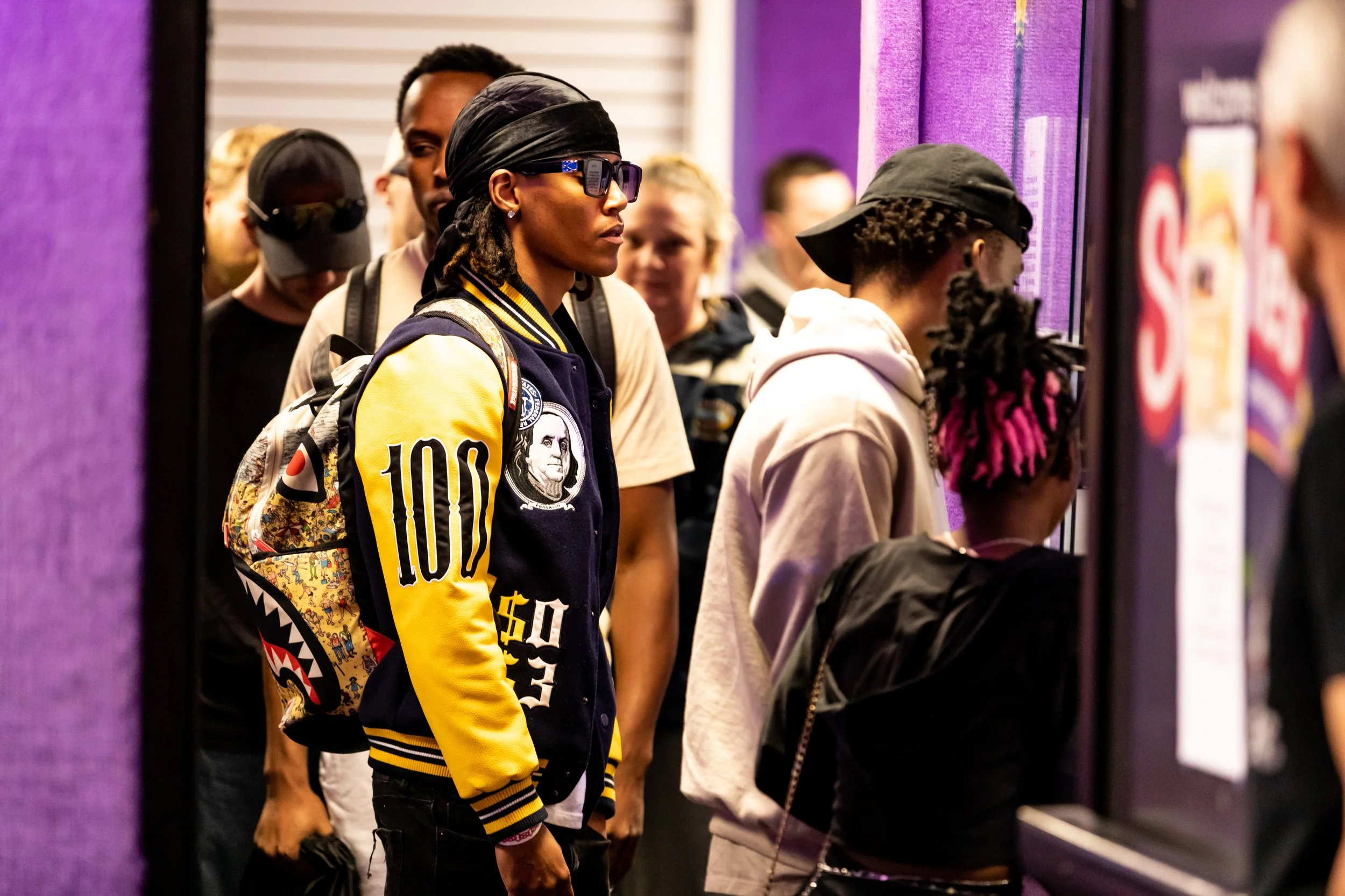 A group of young people waiting in line at an indoor event, with some looking at a display or screen. Purple walls surround them.
