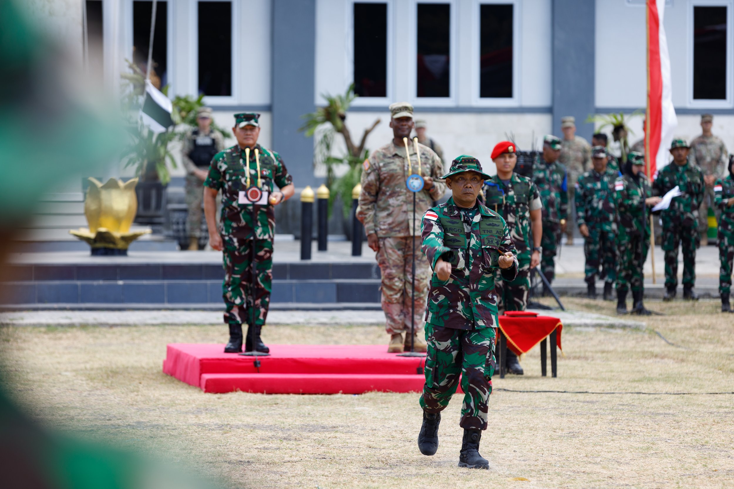 Military officials in camouflage uniforms participating in a formal outdoor ceremony, with some standing on a red carpeted platform and others standing in formation behind them, in front of a modern building.