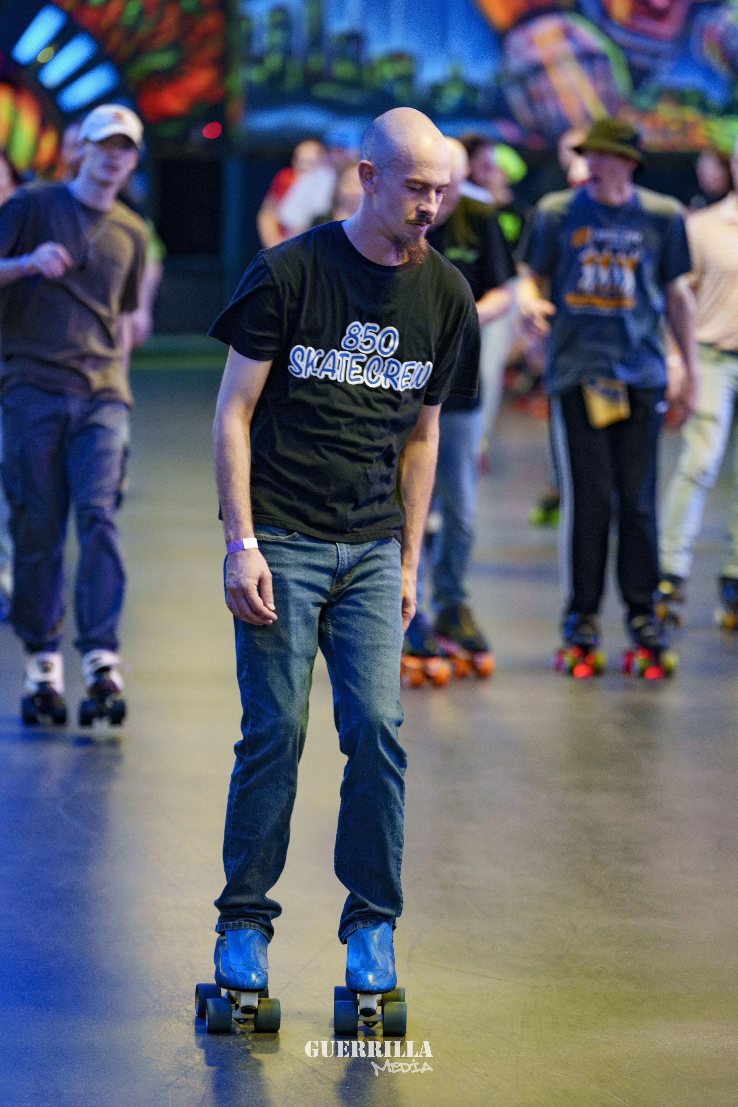 Man with a shaved head and goatee riding a roller skate at an indoor roller skating rink, wearing a black t-shirt that says '850 Skate Crew', with other skaters in the background.