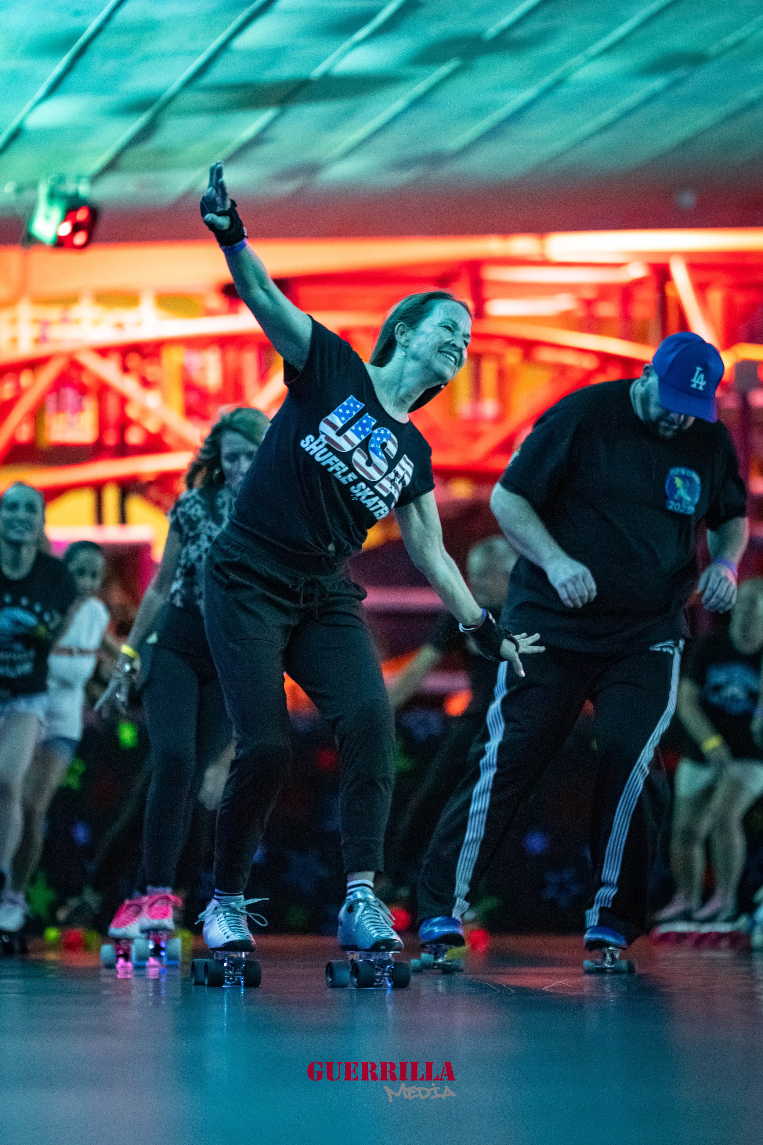 A woman rollerskating indoors with a big smile, raising her right arm in celebration, surrounded by others also skating. Colorful lights and an amusement park ride are visible in the background.
