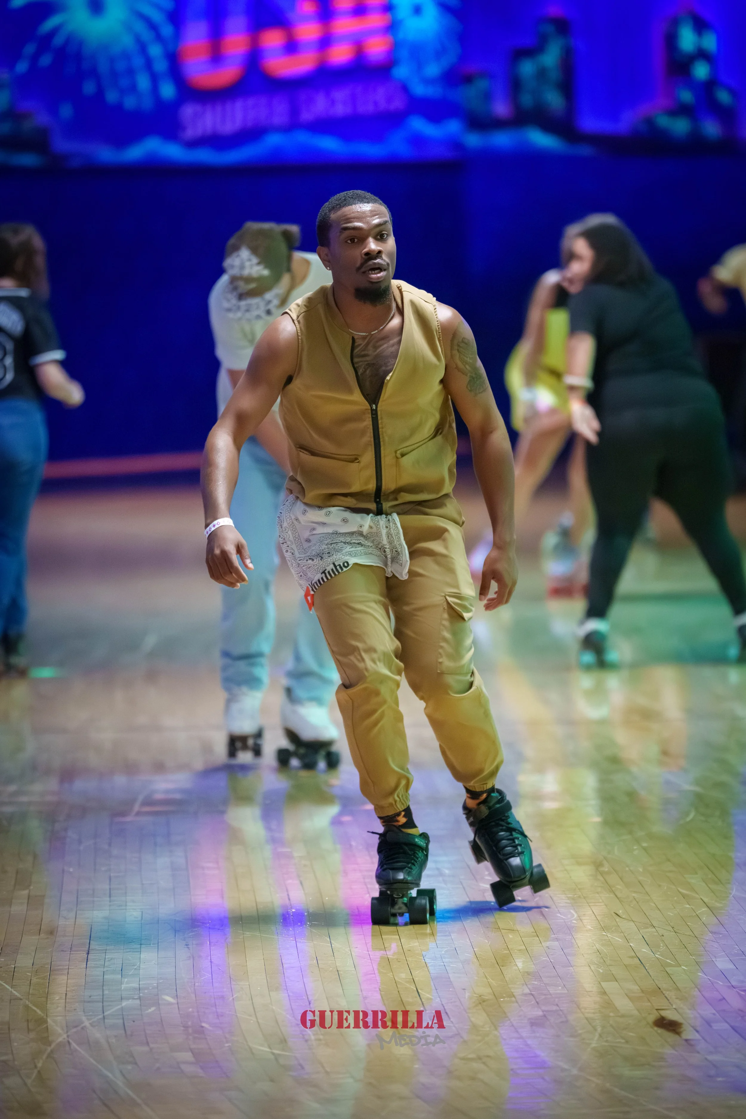 A man in tan outfit roller skating on a wooden floor at an indoor roller skating rink with colorful lights and other people in the background.