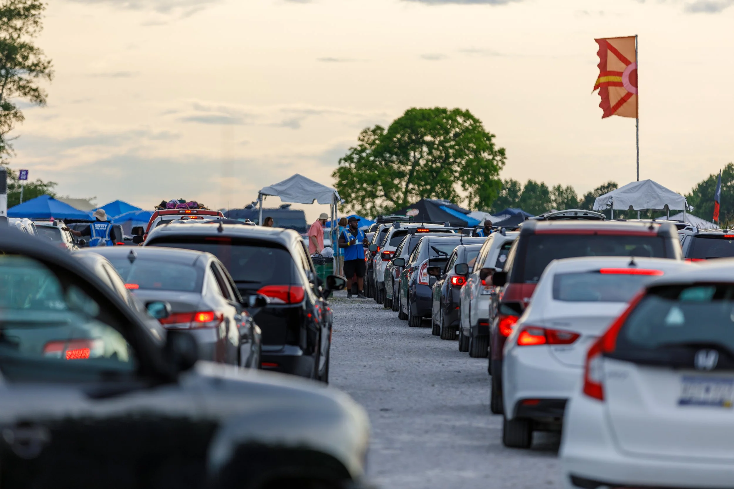 A long line of parked cars extending into the distance at an outdoor event with tents and flags in the background, under a cloudy sky.